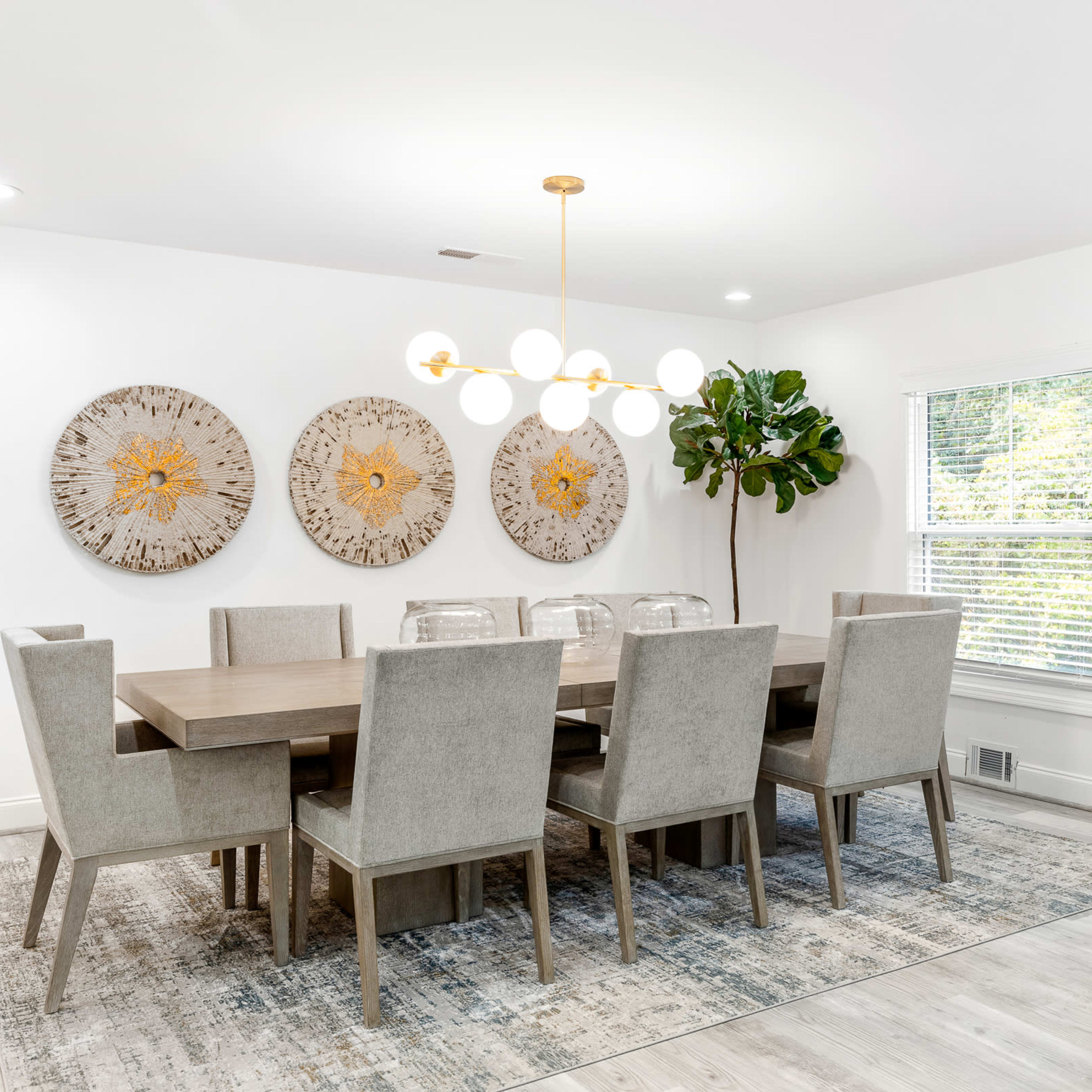 A modern dining room features a long wooden table surrounded by upholstered chairs, with three decorative wall hangings and a bookshelf against the wall.