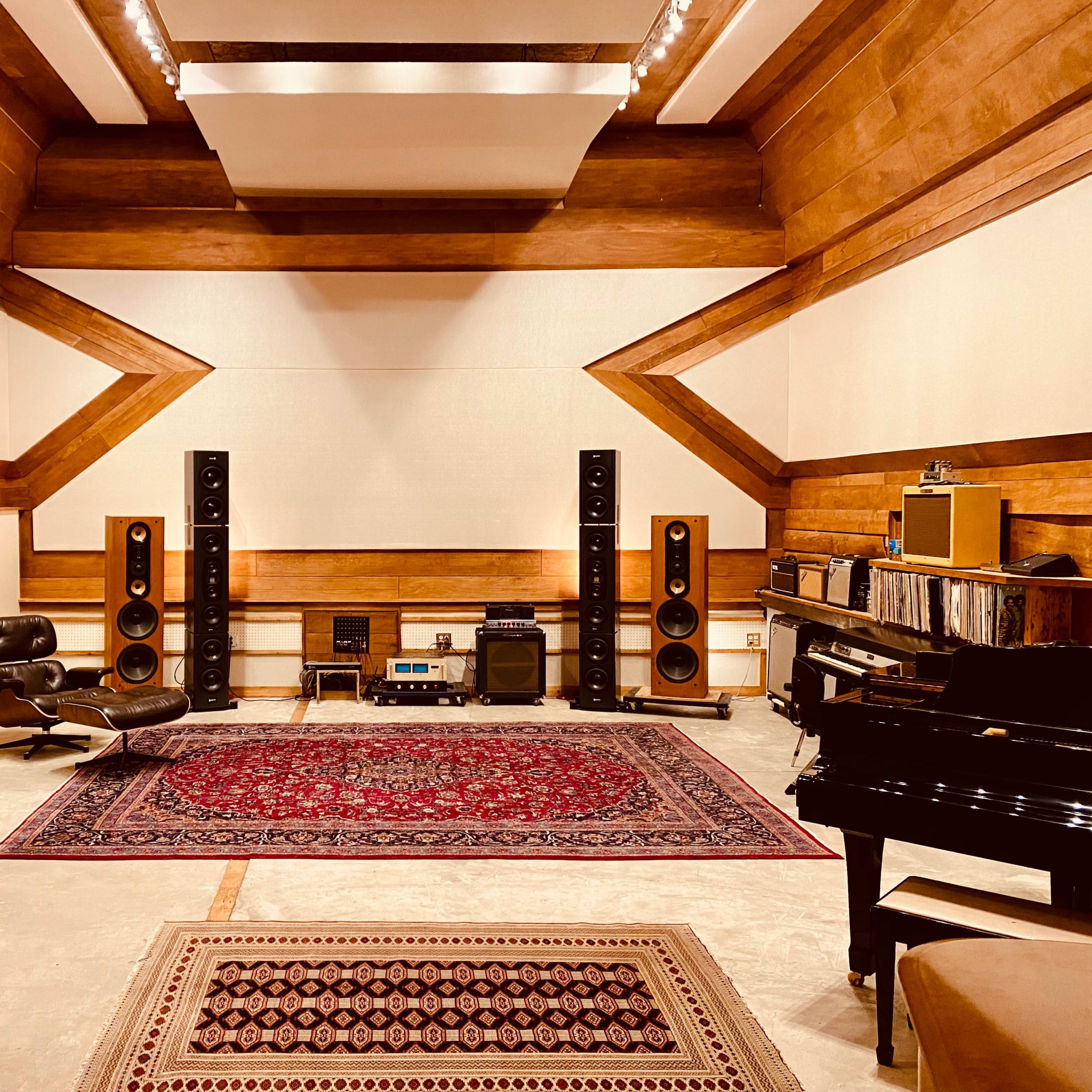 A spacious music studio features vinyl records on shelves, large speakers on either side, a grand piano, and a patterned rug on the floor.