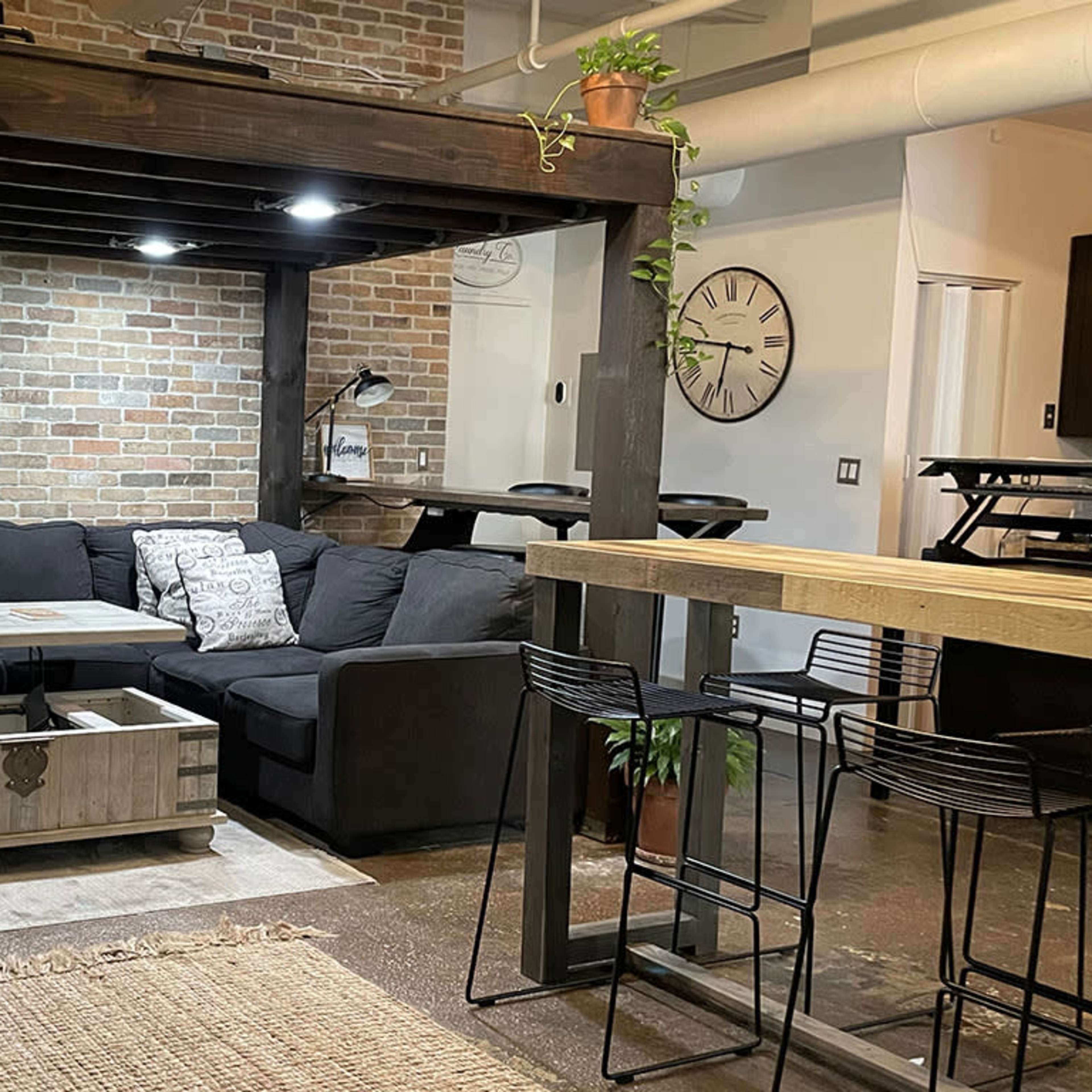 A modern living space with a black sectional sofa, a wooden coffee table, and a brick wall, along with a dining area featuring high stools and a large wooden table.