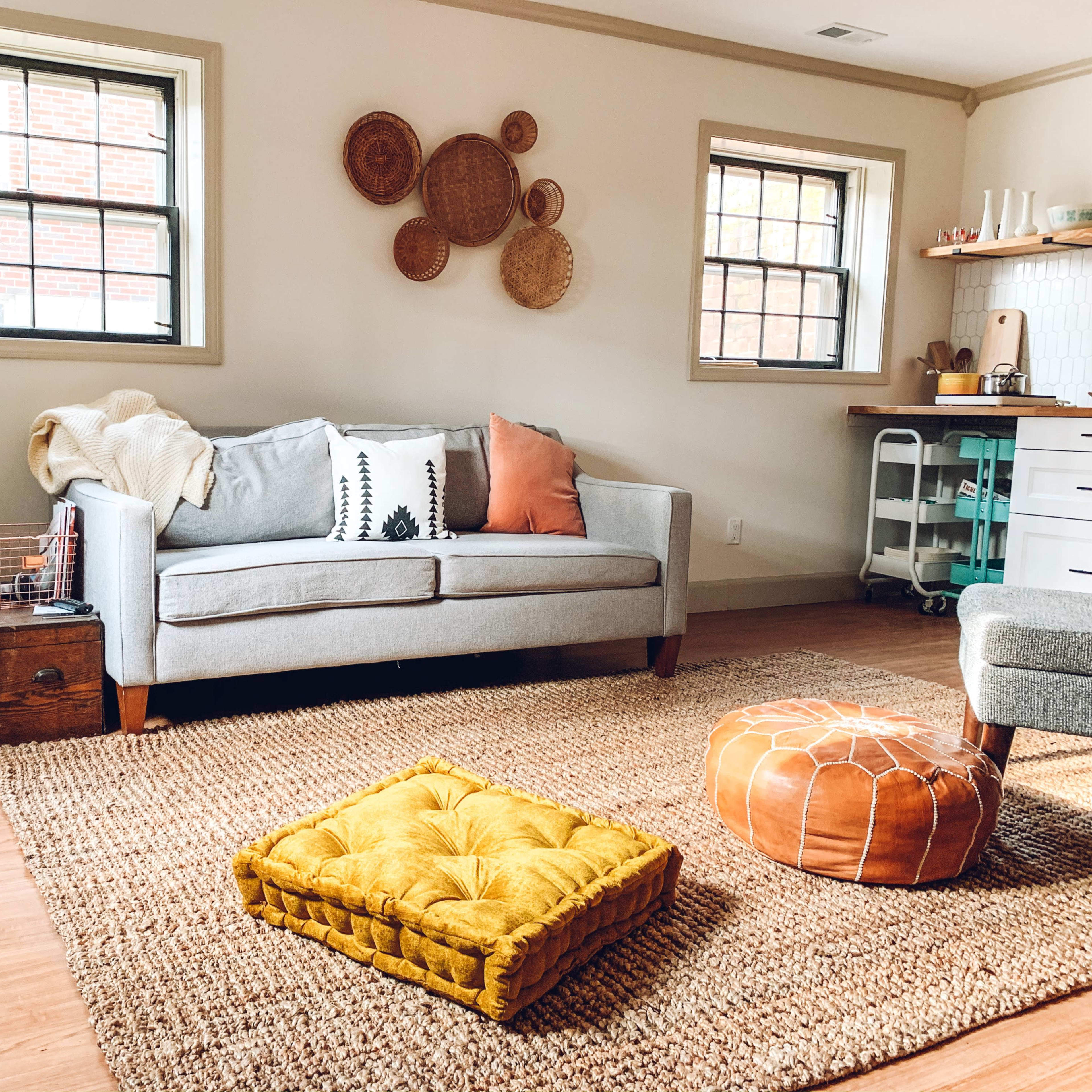 The scene features a cozy living room with a gray sofa, colorful cushions, a round pouf, and decorative wall hangings, set on a woven rug with a potted plant nearby.