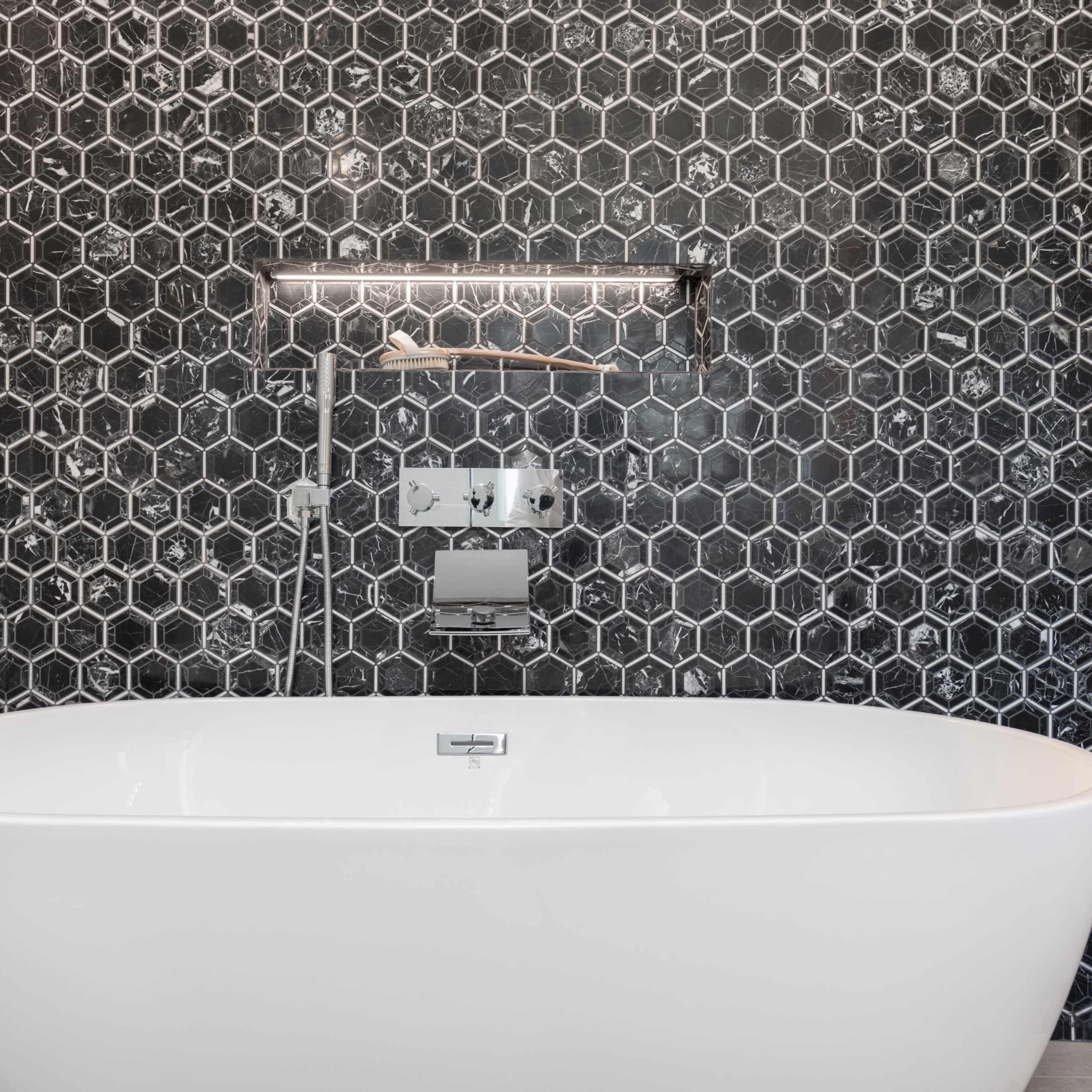 A freestanding white bathtub is positioned against a hexagonal patterned black and gray tiled wall, illuminated by a modern wall-mounted light fixture.