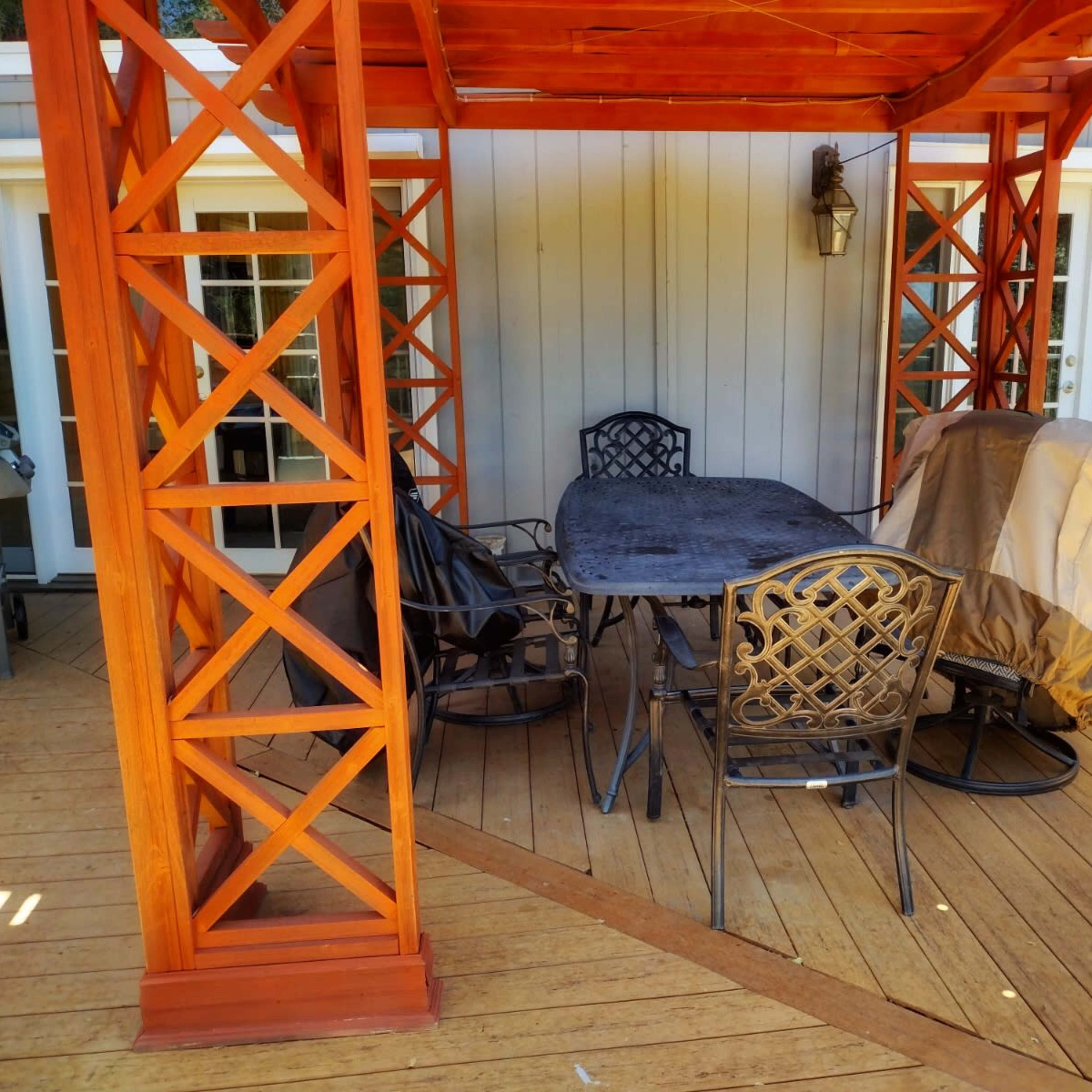 A wooden gazebo with a table and chairs is set on a deck, accompanied by a covered grill.