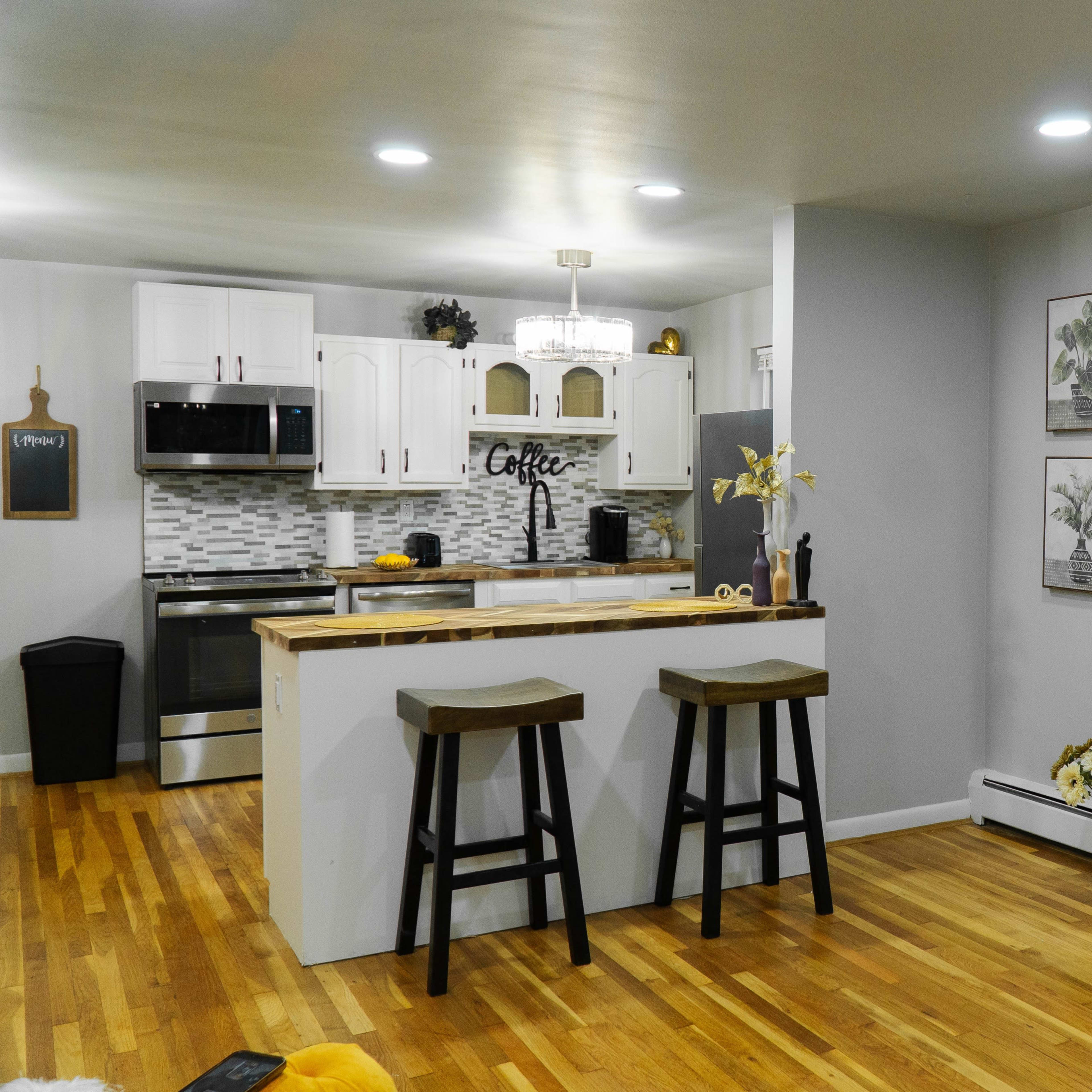 A modern kitchen and living area featuring white cabinetry, a wooden island with two stools, and an electric fireplace with a wood stack beneath a mounted TV.