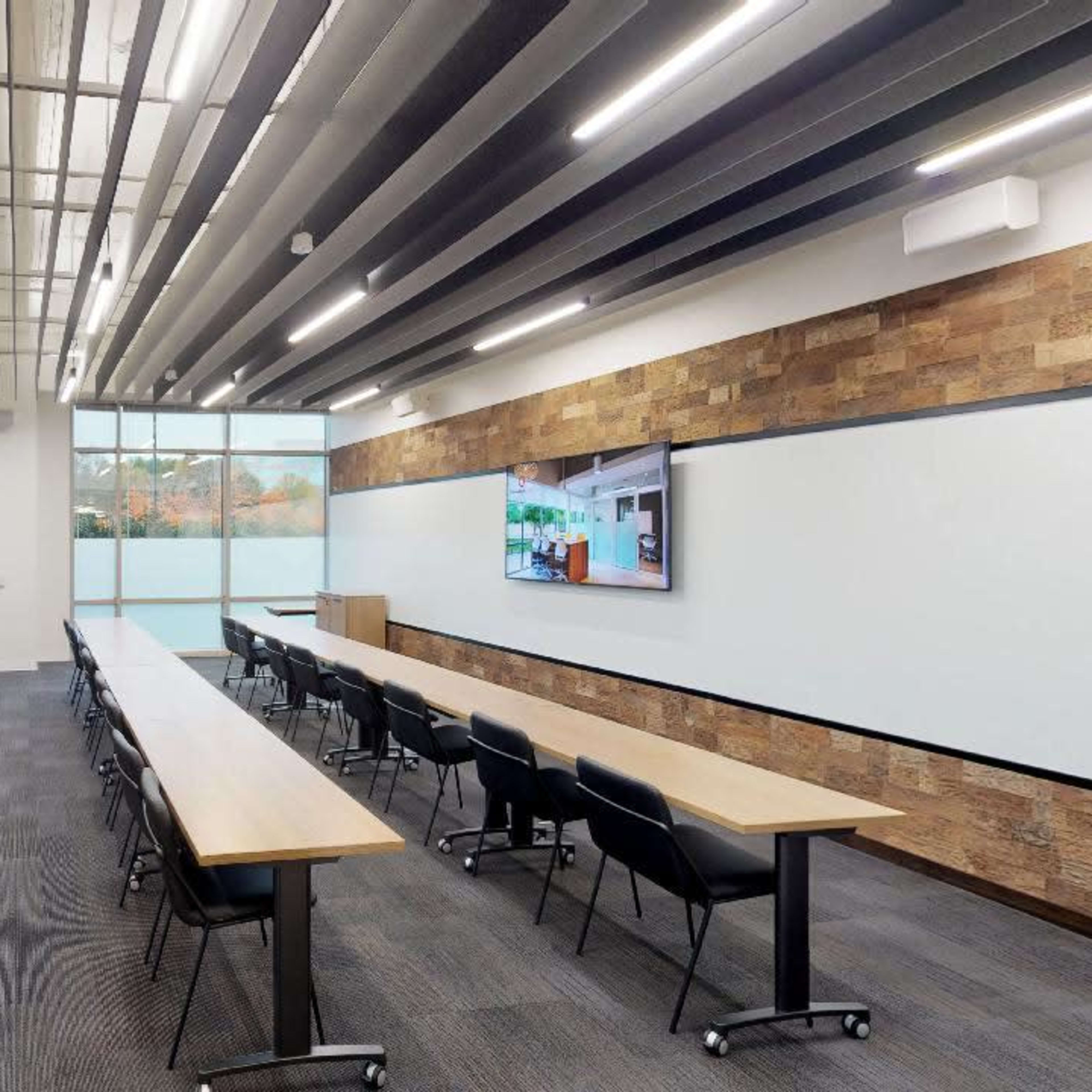 A modern conference room with long tables, chairs, large windows, and a screen displaying digital content.