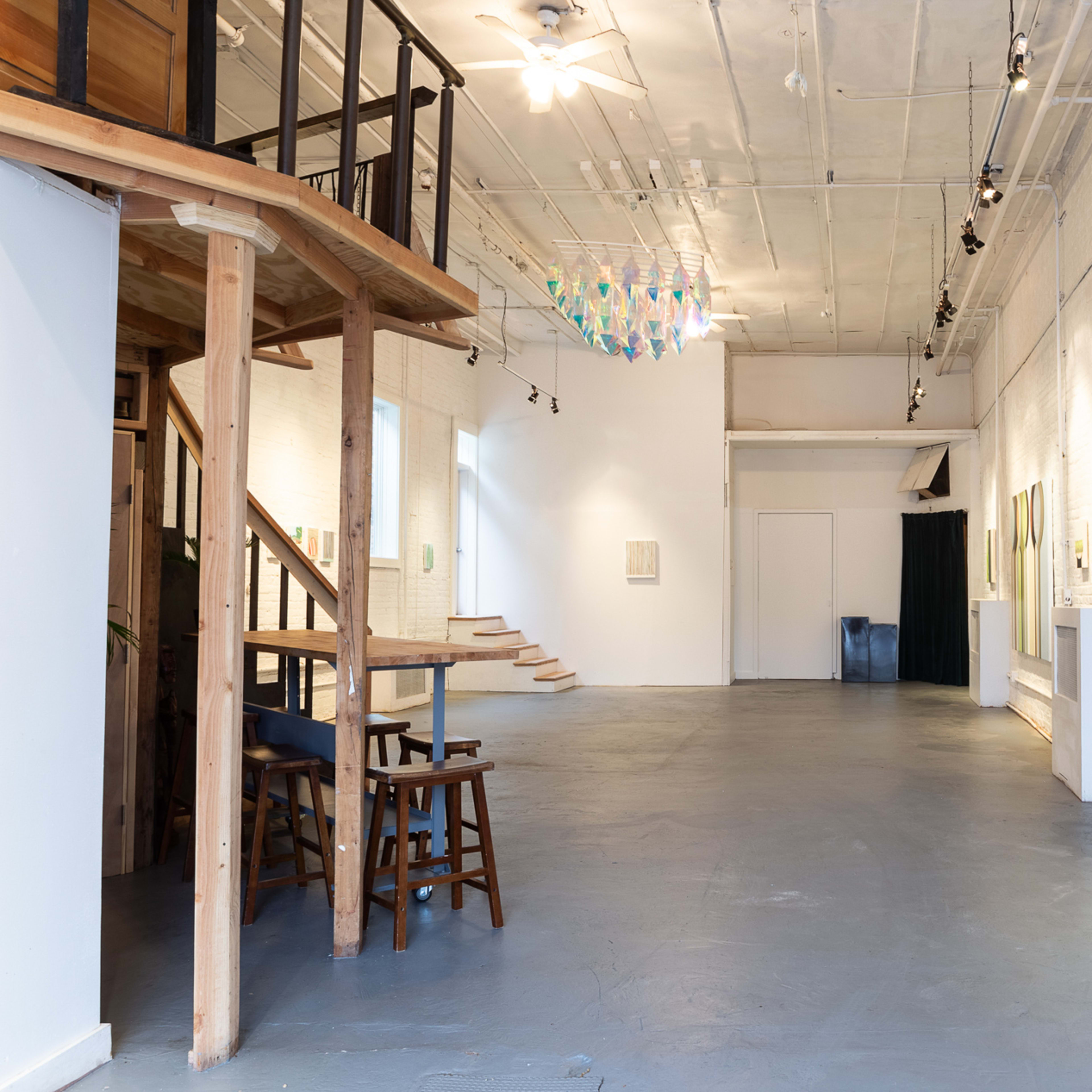 A spacious art gallery with wooden stairs on the left, various artworks on the walls, and colorful hanging sculptures above.