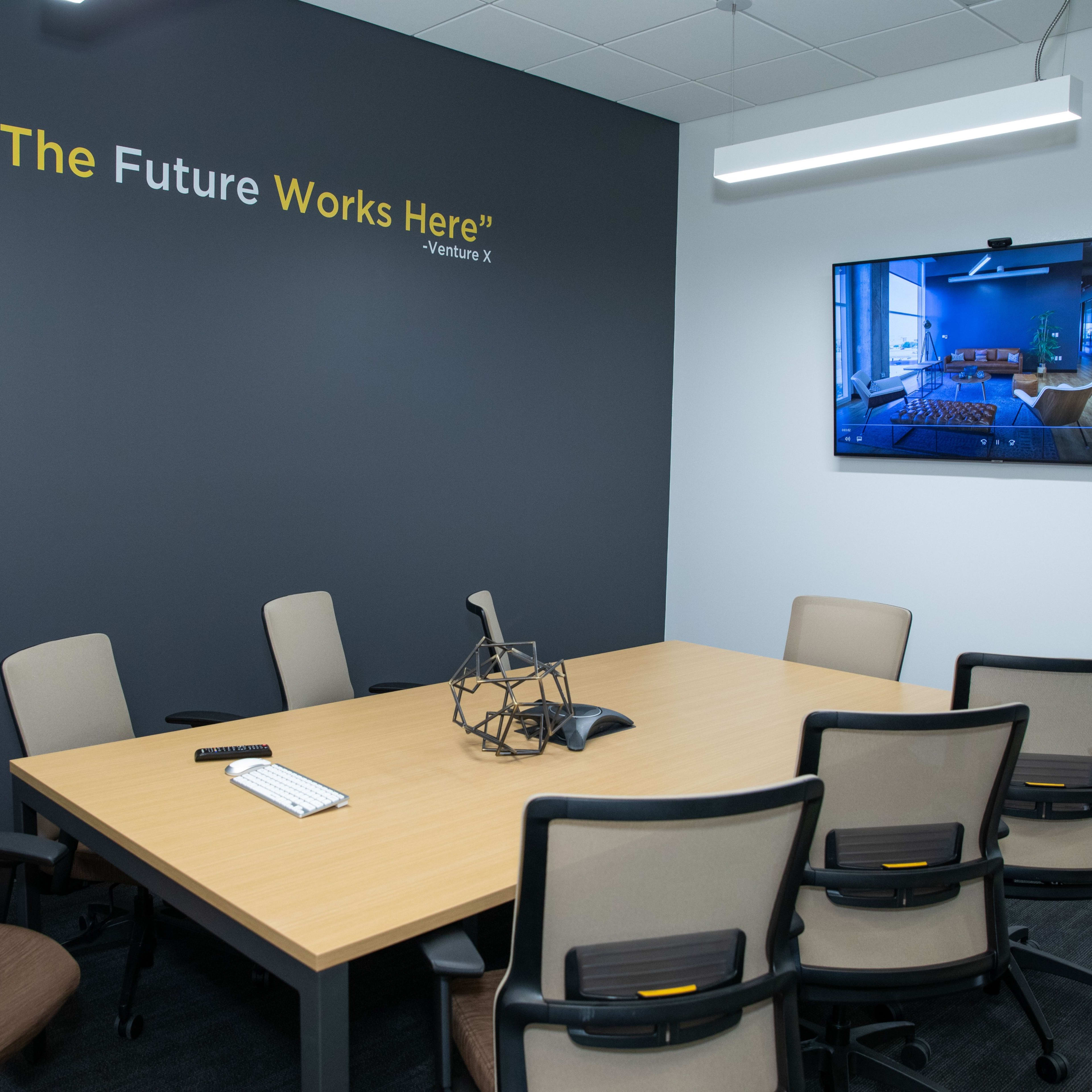 A conference room features a table surrounded by chairs, with a large screen displaying an image on the wall next to a quote about the future.