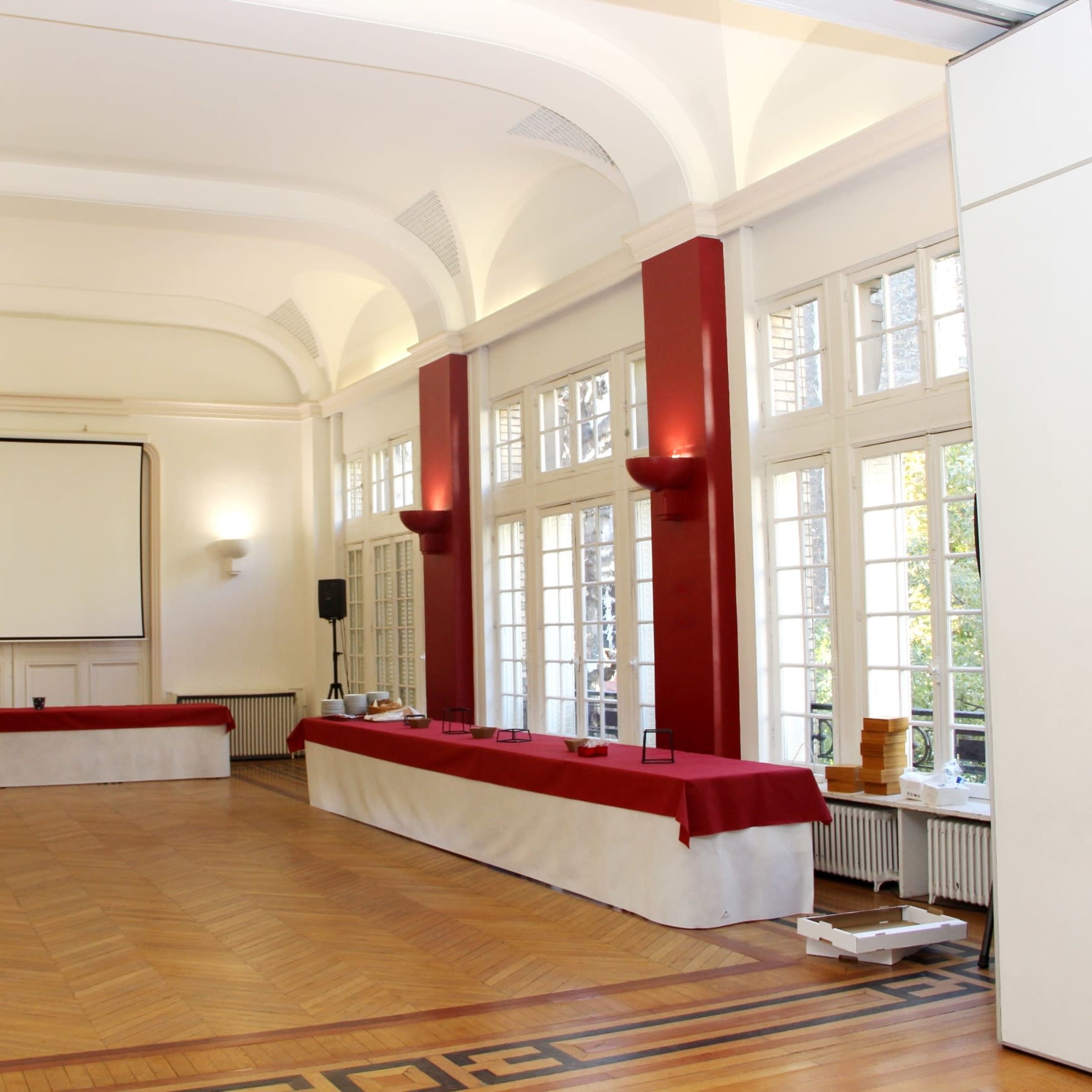 A spacious room features a large screen and a long table covered with a red cloth, flanked by large windows letting in natural light.