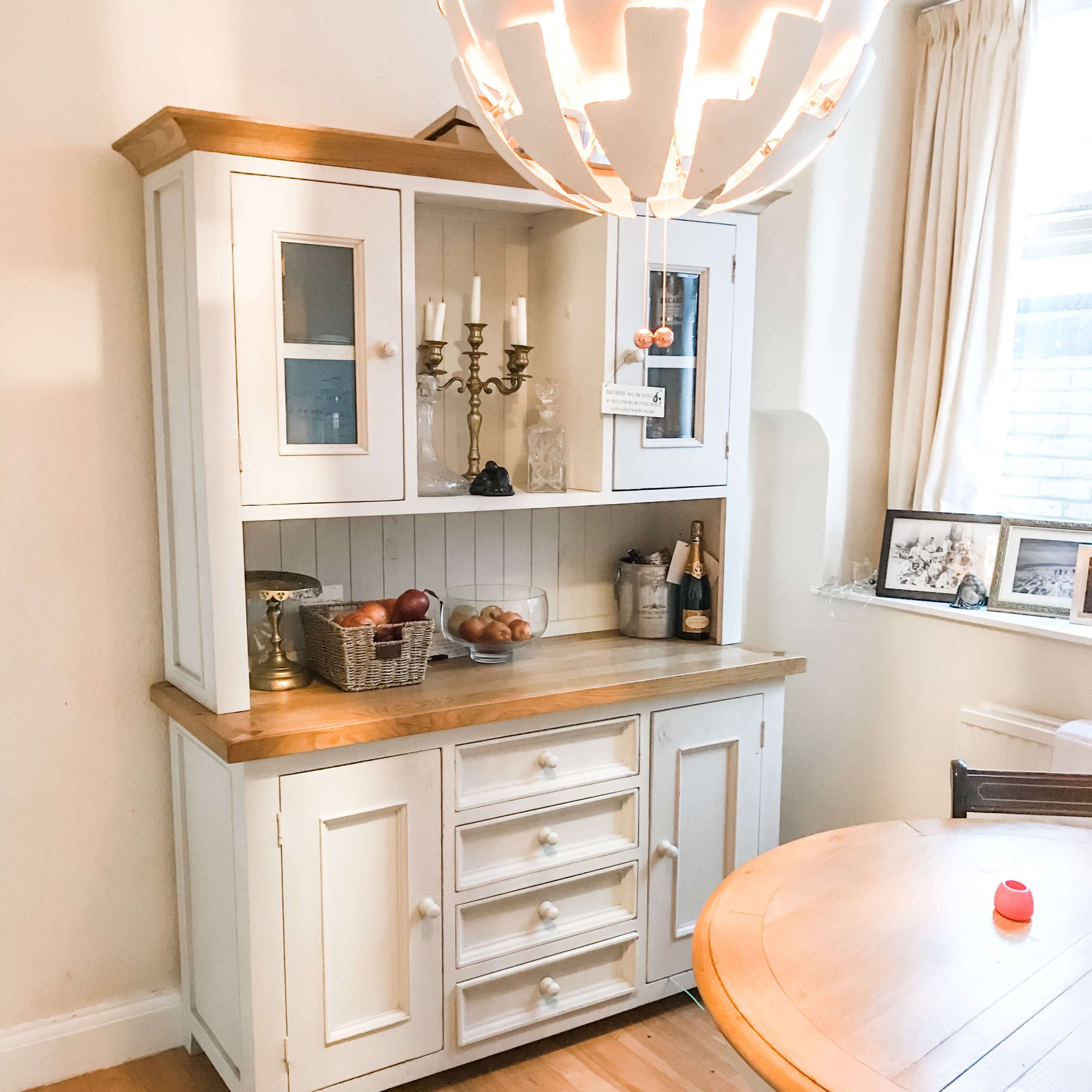 A wooden sideboard with glass doors and a countertop displays a basket of fruit and two bottles of champagne near a round dining table.