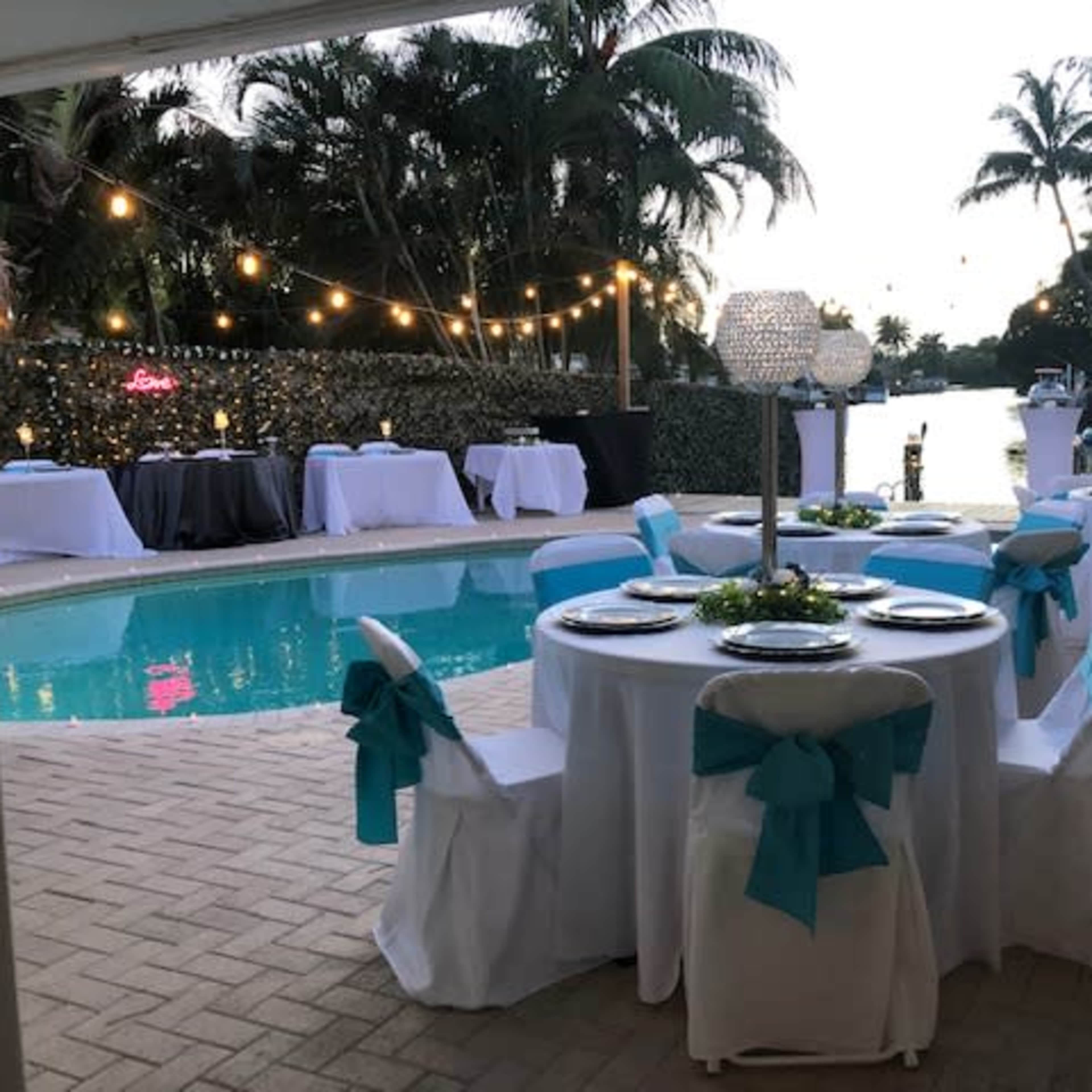 A poolside event space is set up with tables draped in white linens and adorned with turquoise ribbons, surrounded by tropical plants and string lights.