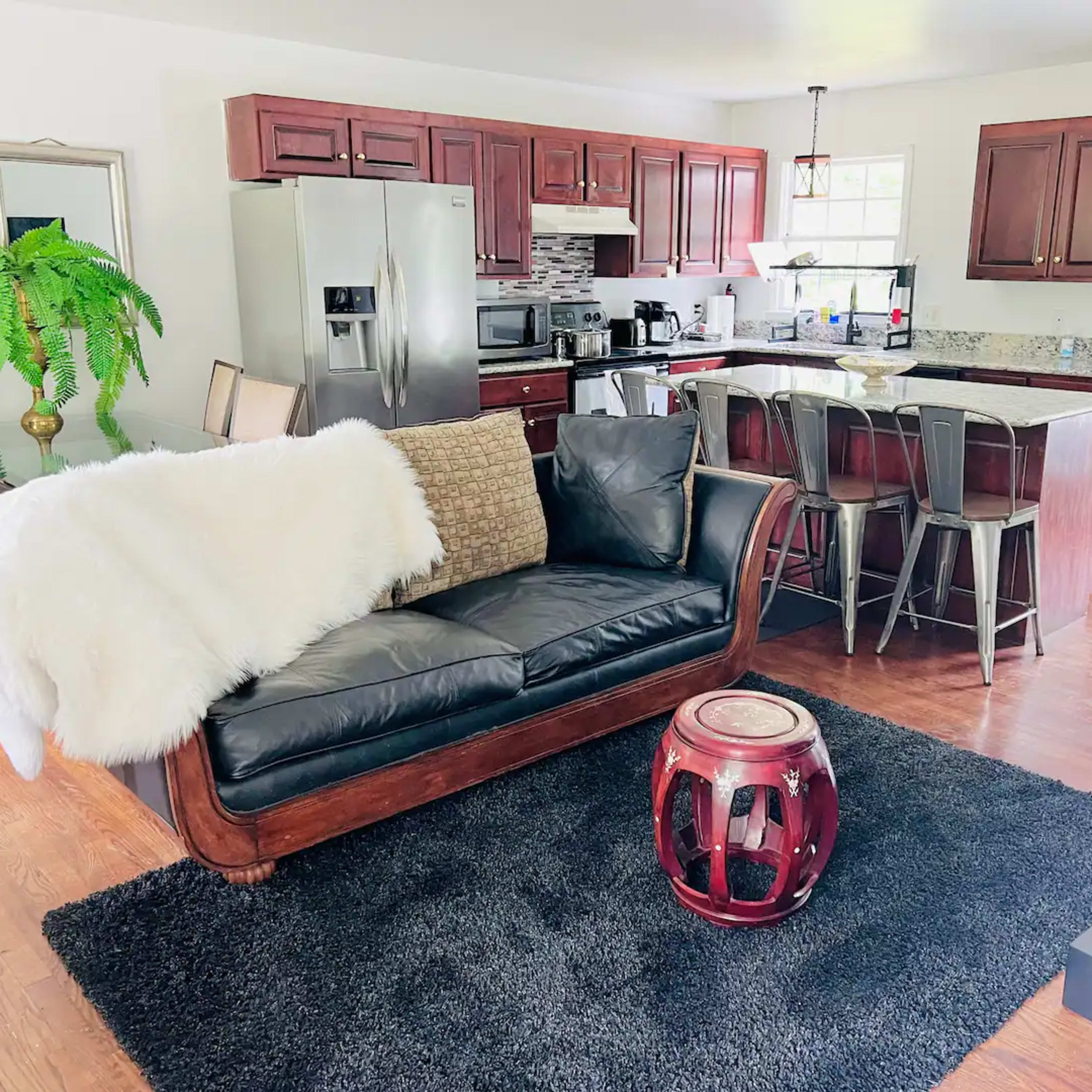 A modern open-plan living area with a black leather sofa, a woven throw, a round red table, and a kitchen area featuring wooden cabinets and stainless steel appliances.