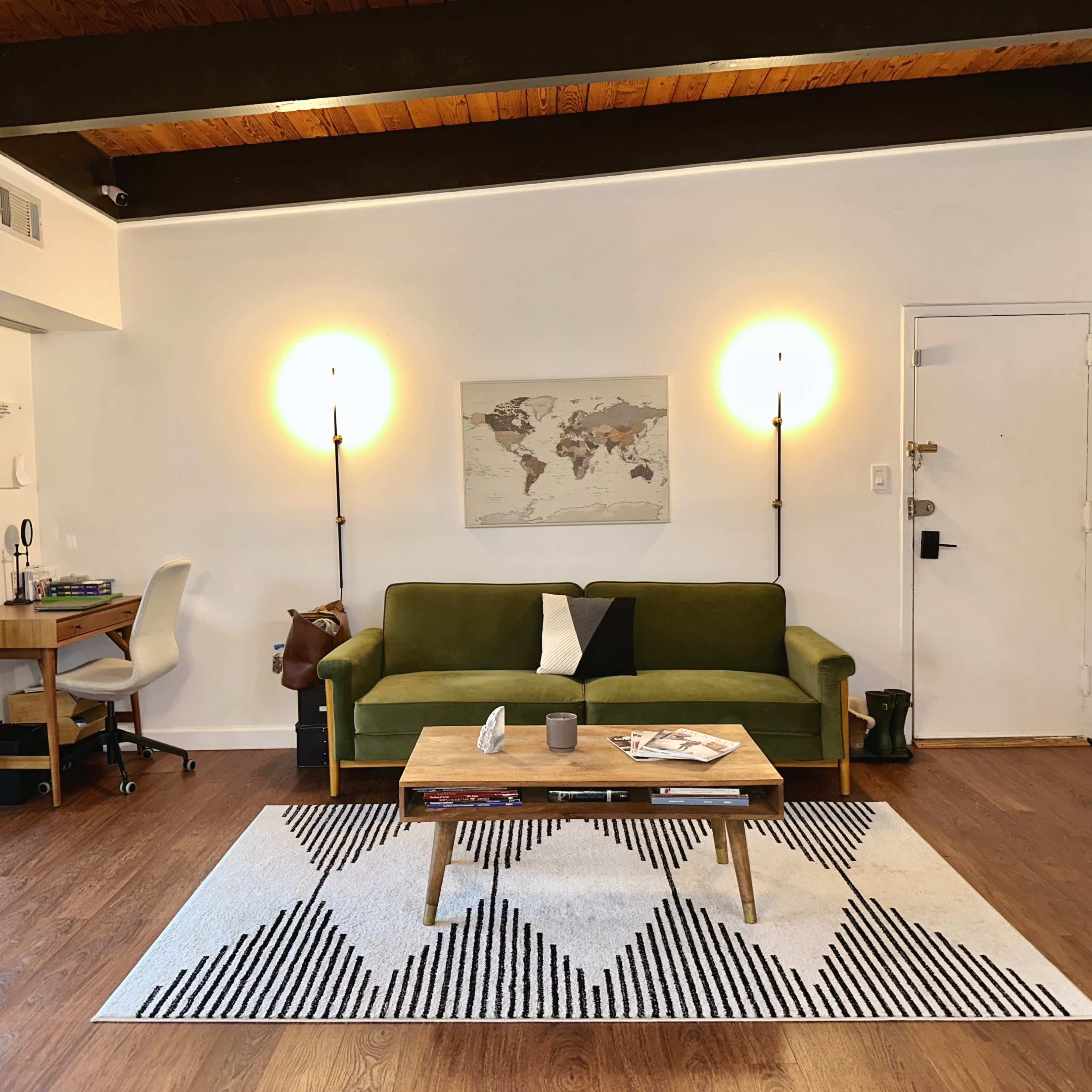 A living room with a green sofa, a wooden coffee table with decorative items, and a desk in the corner beside a large window.