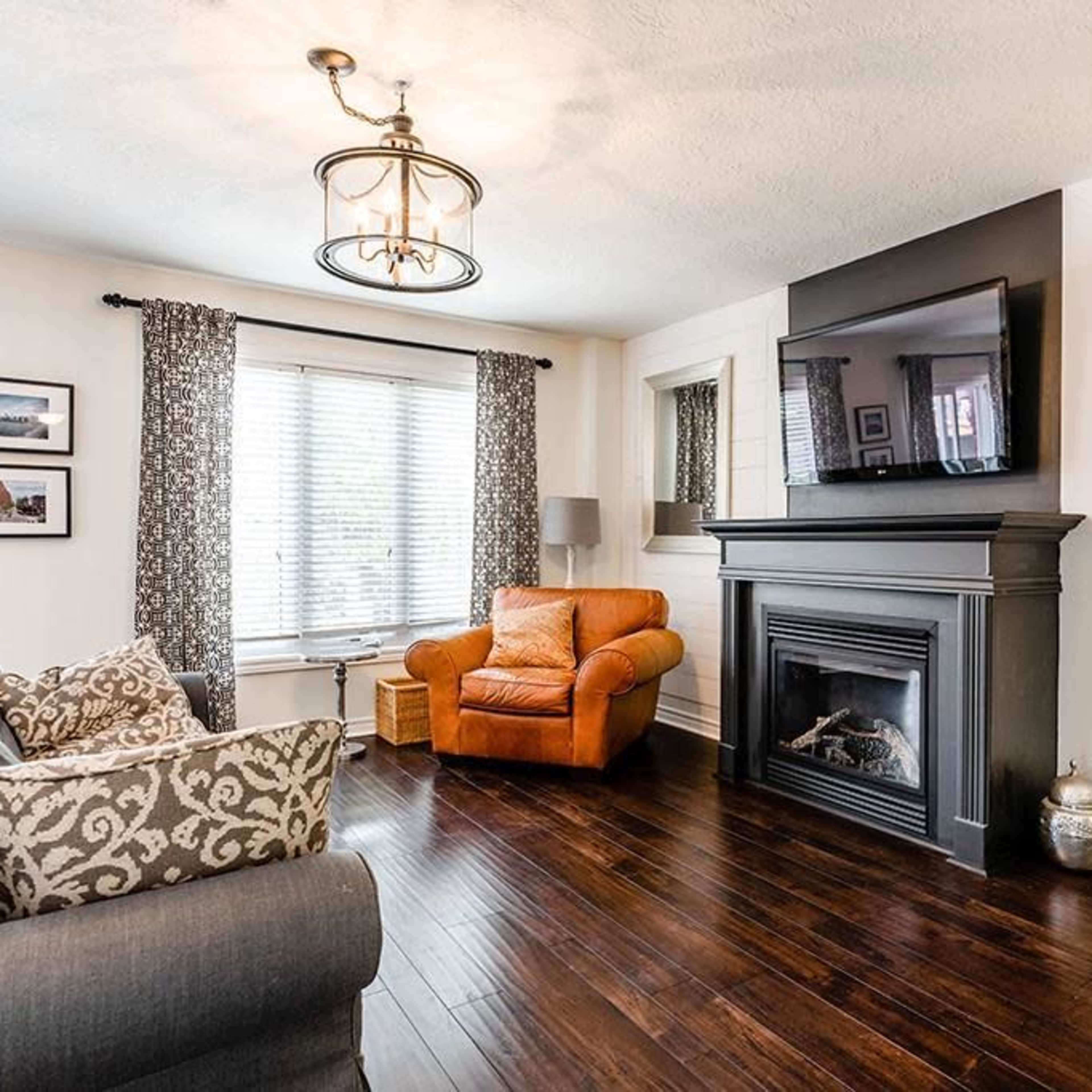The living room features a brown leather armchair, a black fireplace, and a large wall-mounted television, with large windows allowing natural light.