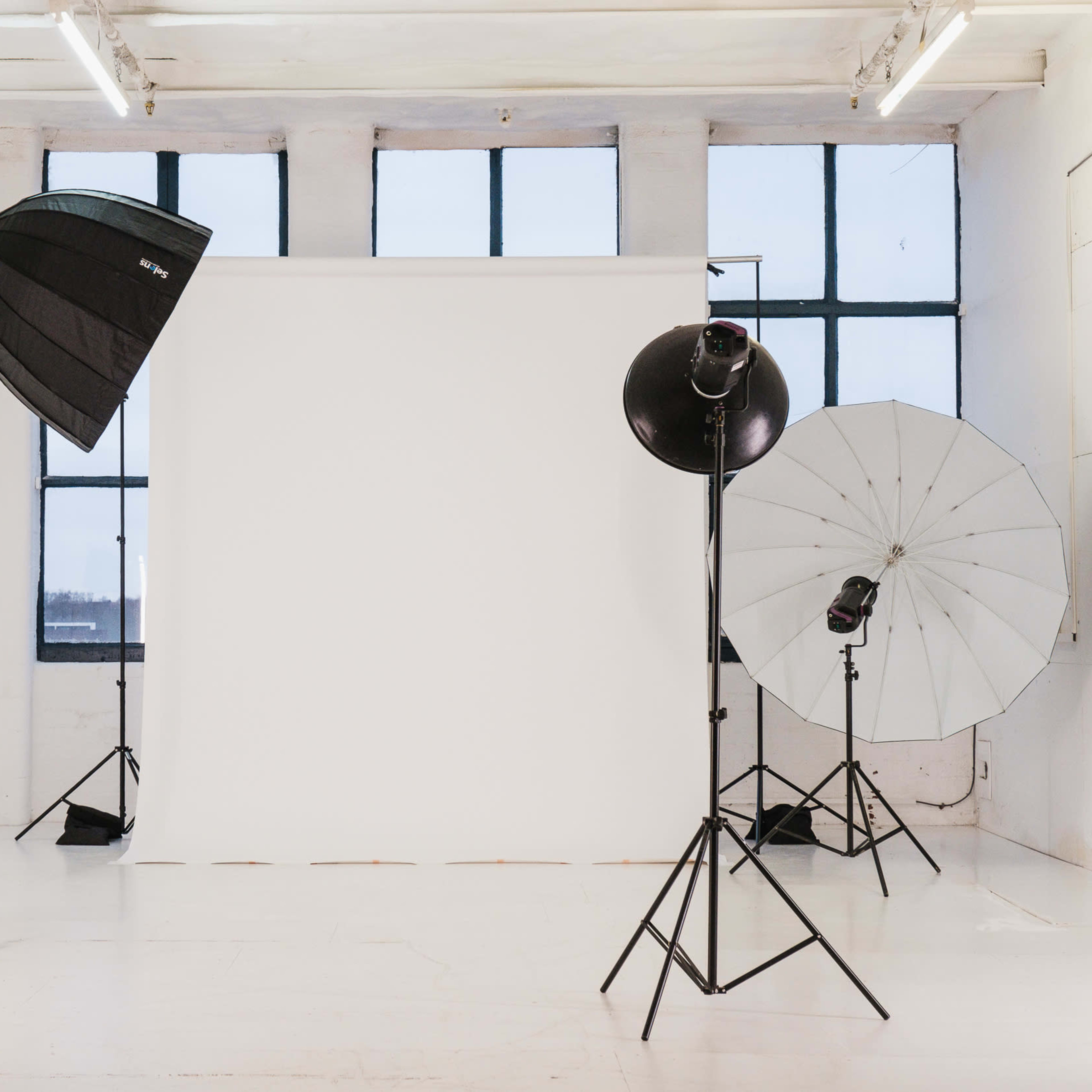 A photography studio features a white backdrop with multiple light sources and equipment arranged around it.