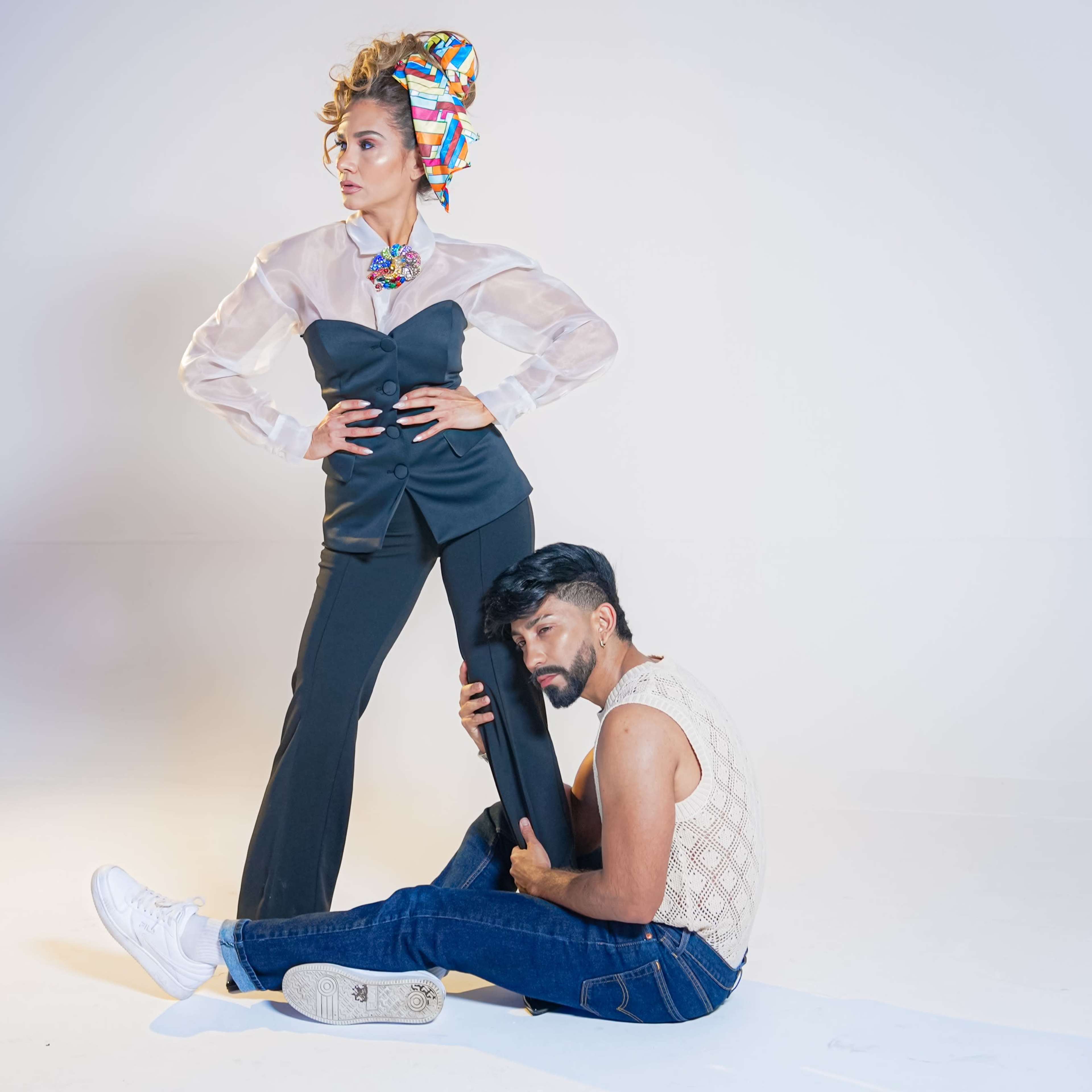 A woman in a sheer blouse and tailored pants stands confidently while a man in a knit top sits on the ground, holding onto her leg.