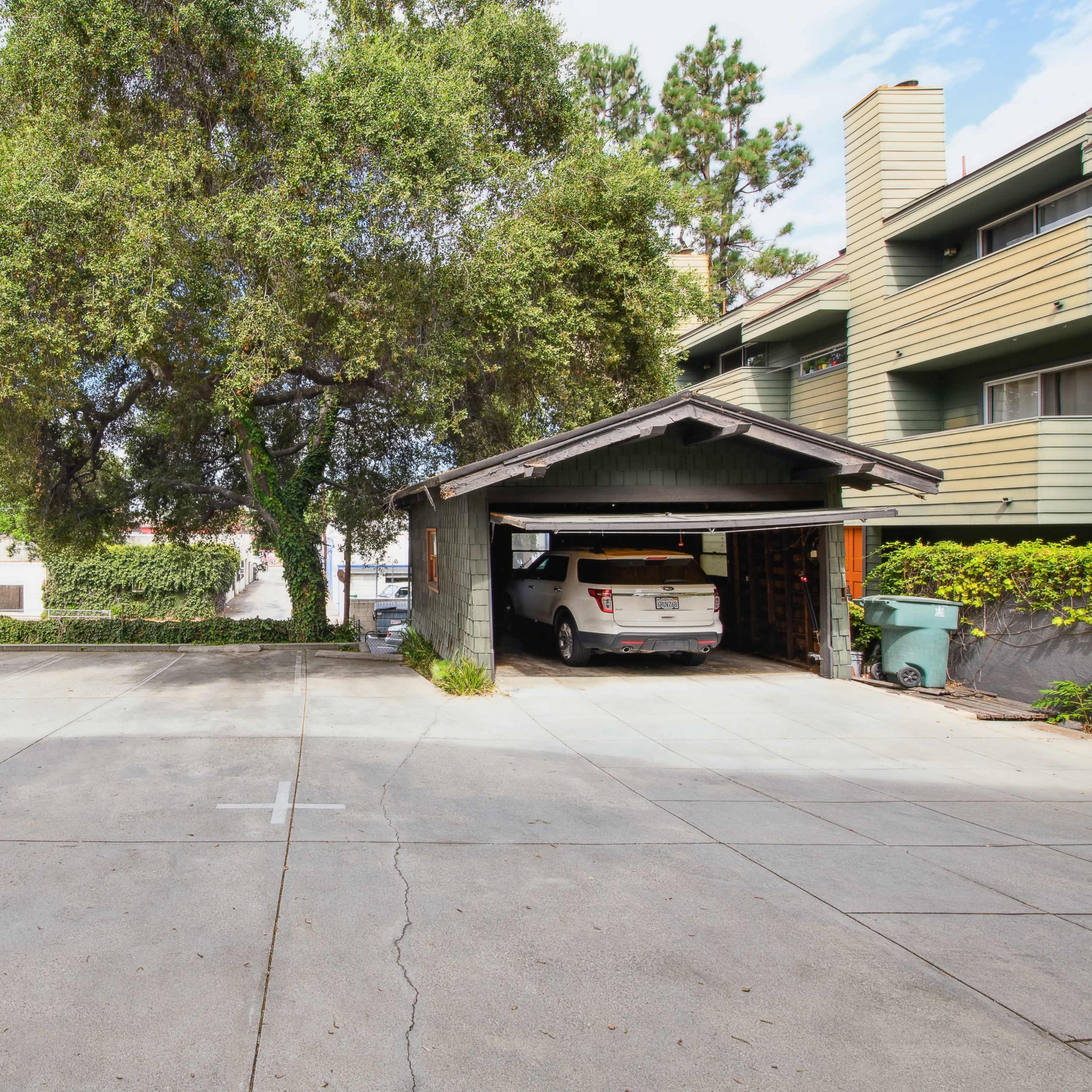 A spacious parking area features a large tree and a car parked under a shaded carport next to a multi-story building.