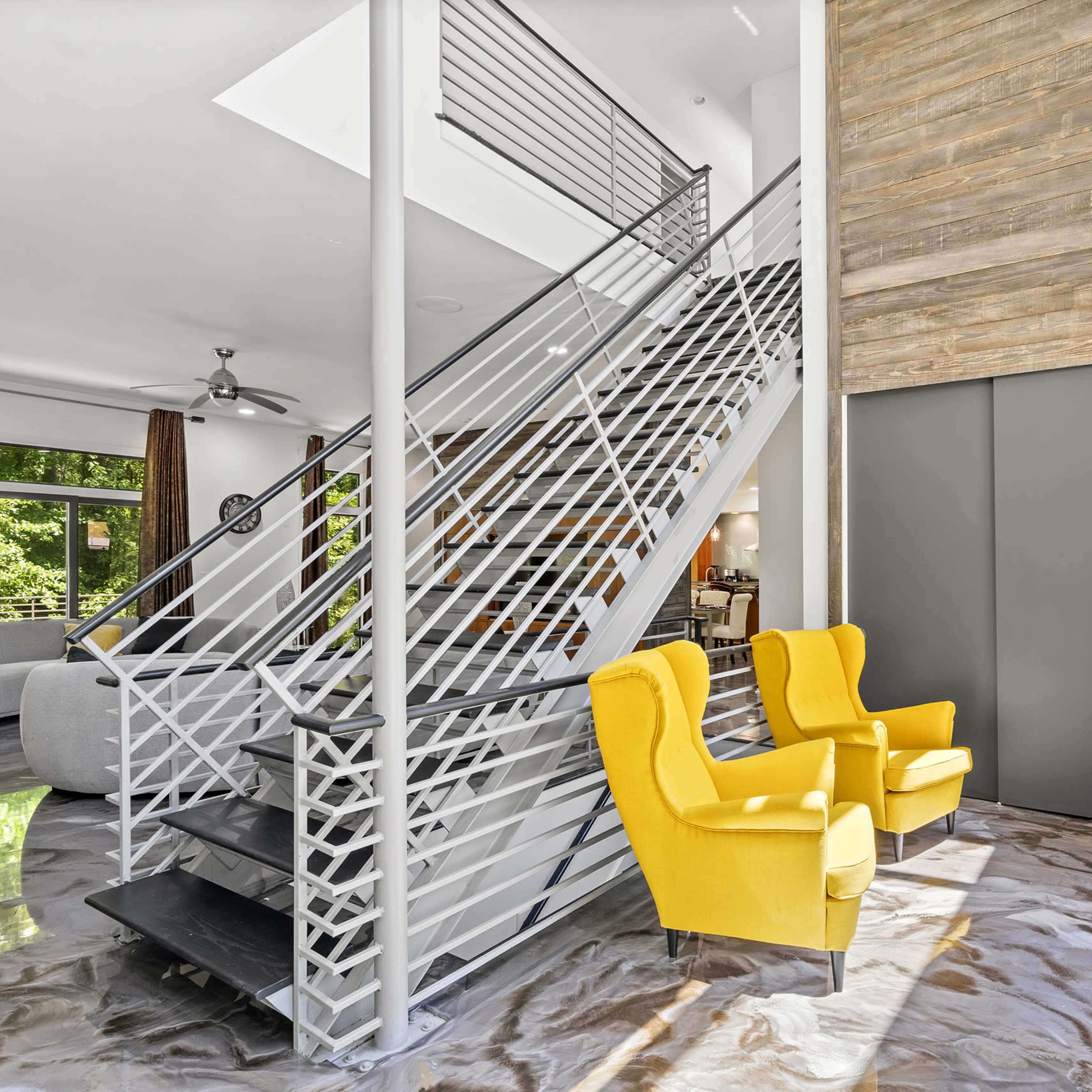 A modern interior featuring a staircase with a metal railing, two yellow armchairs, and large windows that provide natural light.