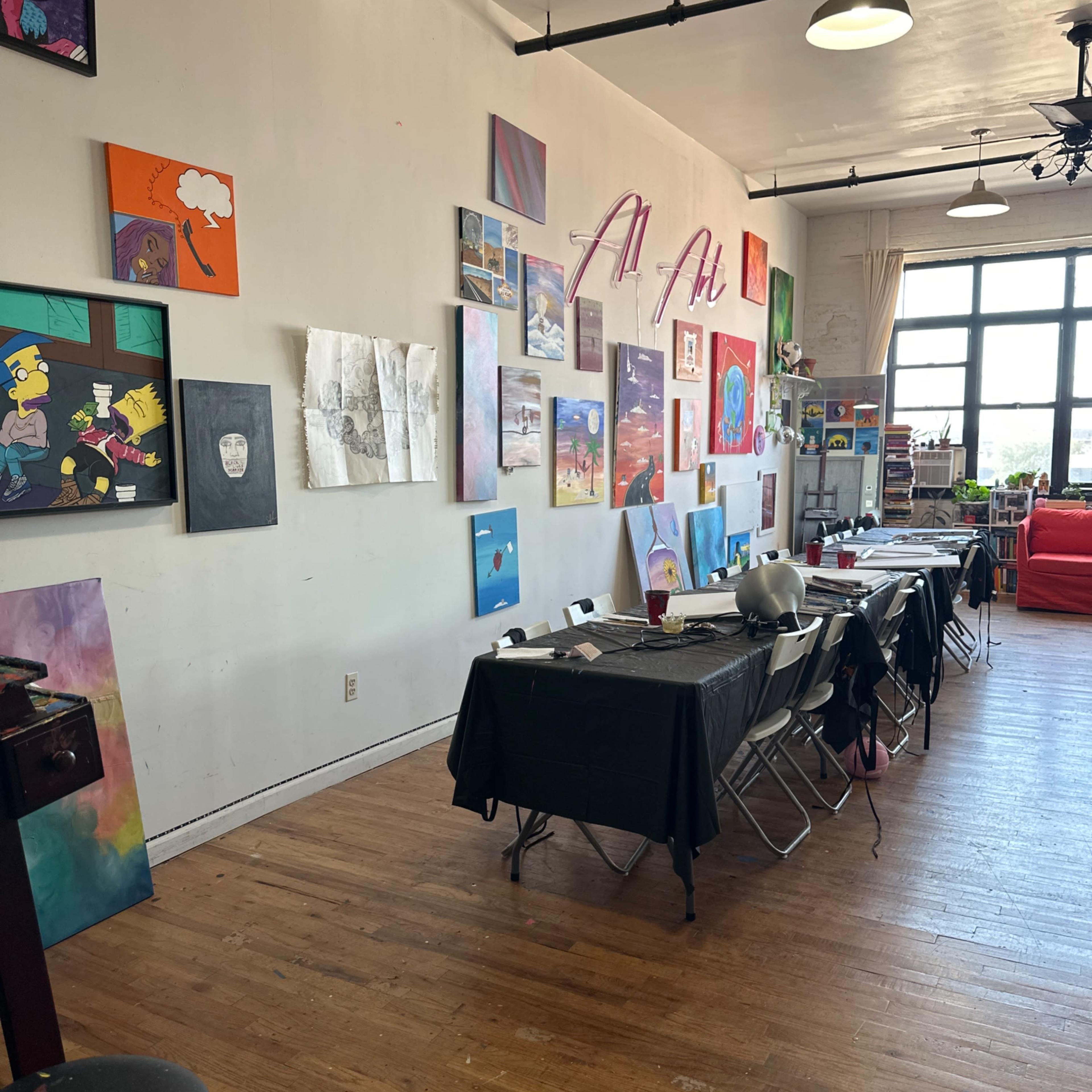 An art studio with a row of tables and chairs on one side, while the walls are adorned with various colorful artworks and posters.