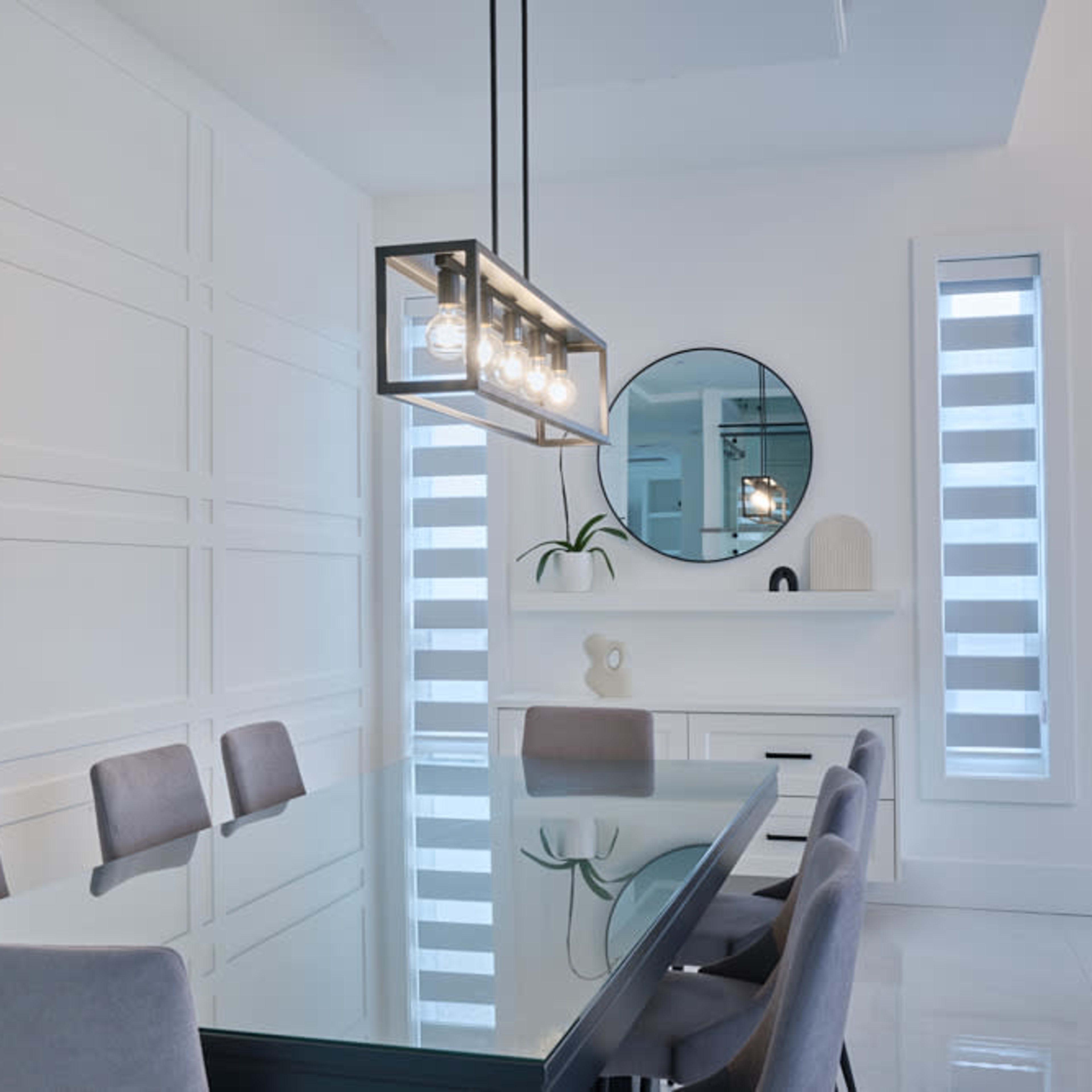 A modern dining room featuring a glass dining table with gray upholstered chairs, a round mirror, and a minimalist lighting fixture.