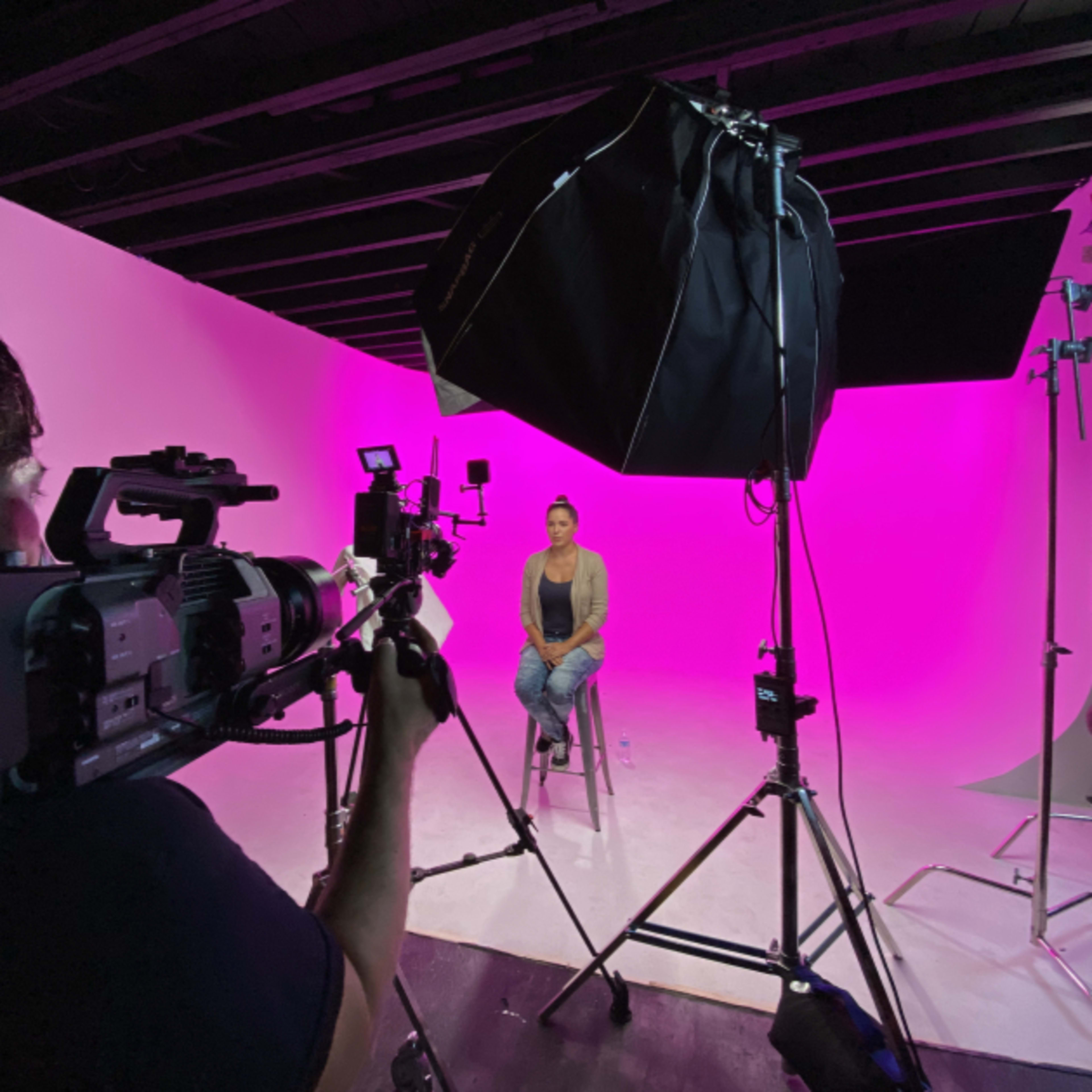 A person sits on a stool in front of a bright pink backdrop while being filmed by a camera crew.