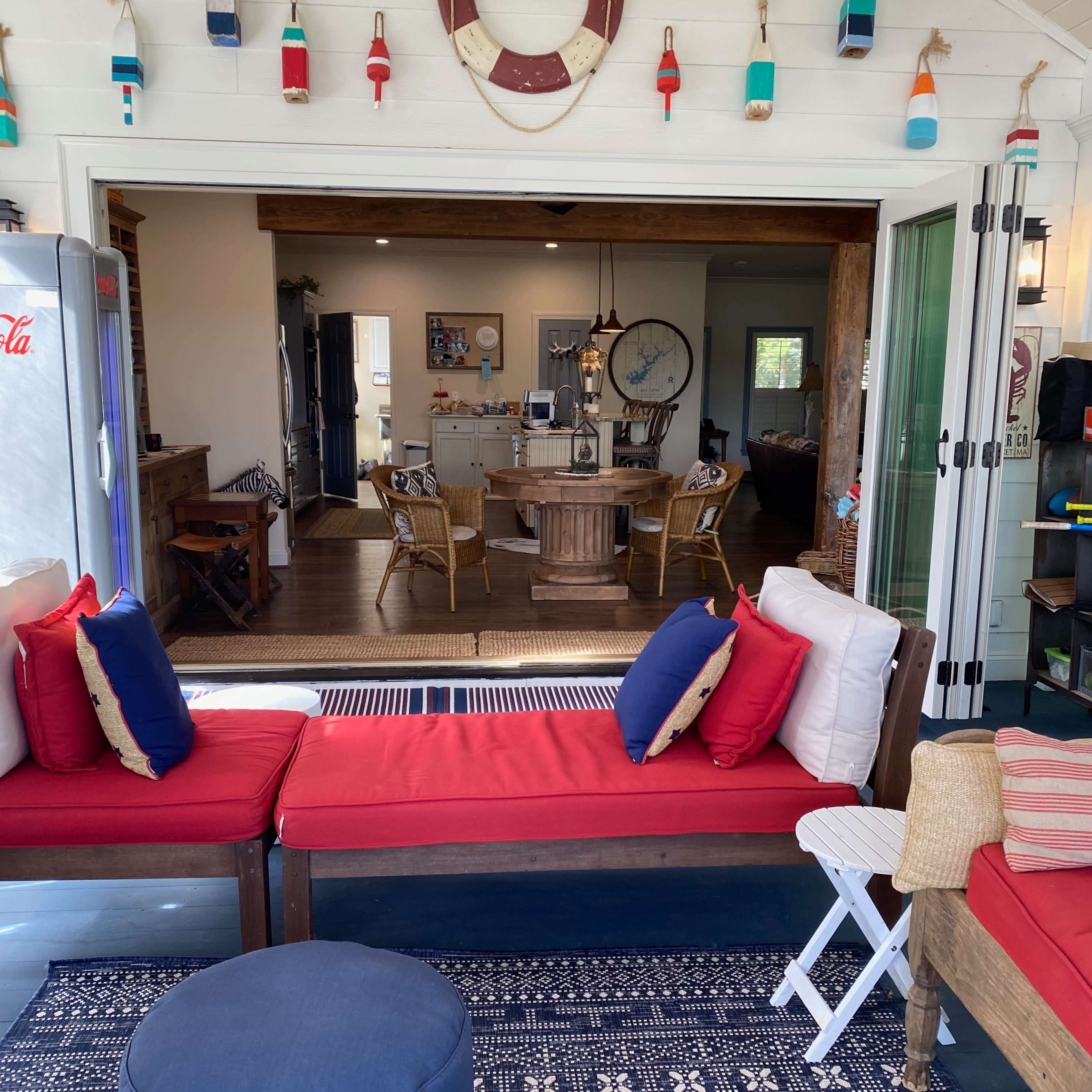 A cozy outdoor seating area with red cushions, a blue pouf, and a view of a well-decorated indoor space through open glass doors.