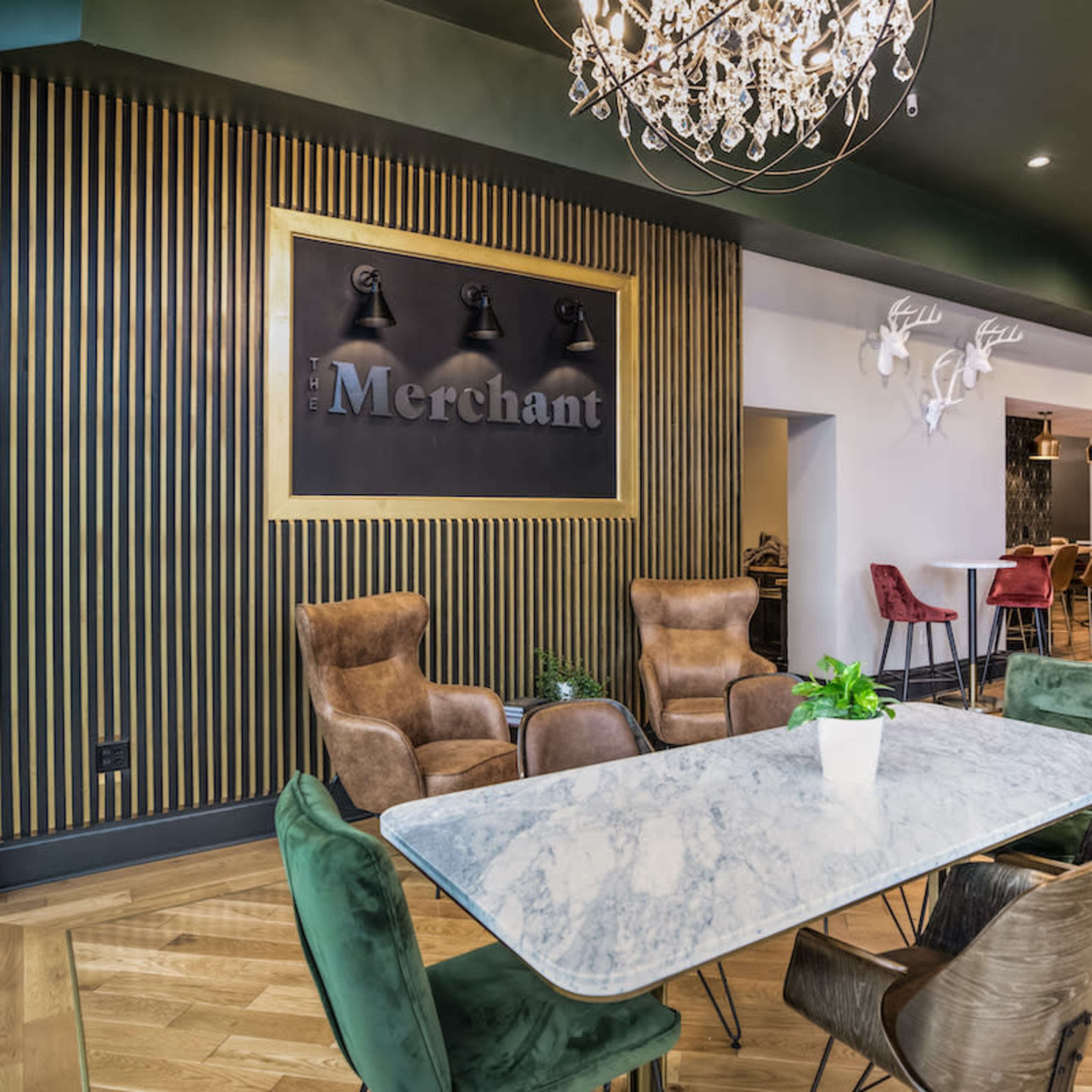 A stylish dining area featuring a large marble table surrounded by upholstered chairs, with a decorative wall displaying the word "Merchant" and accent lighting from chandeliers and wall-mounted fixtures.