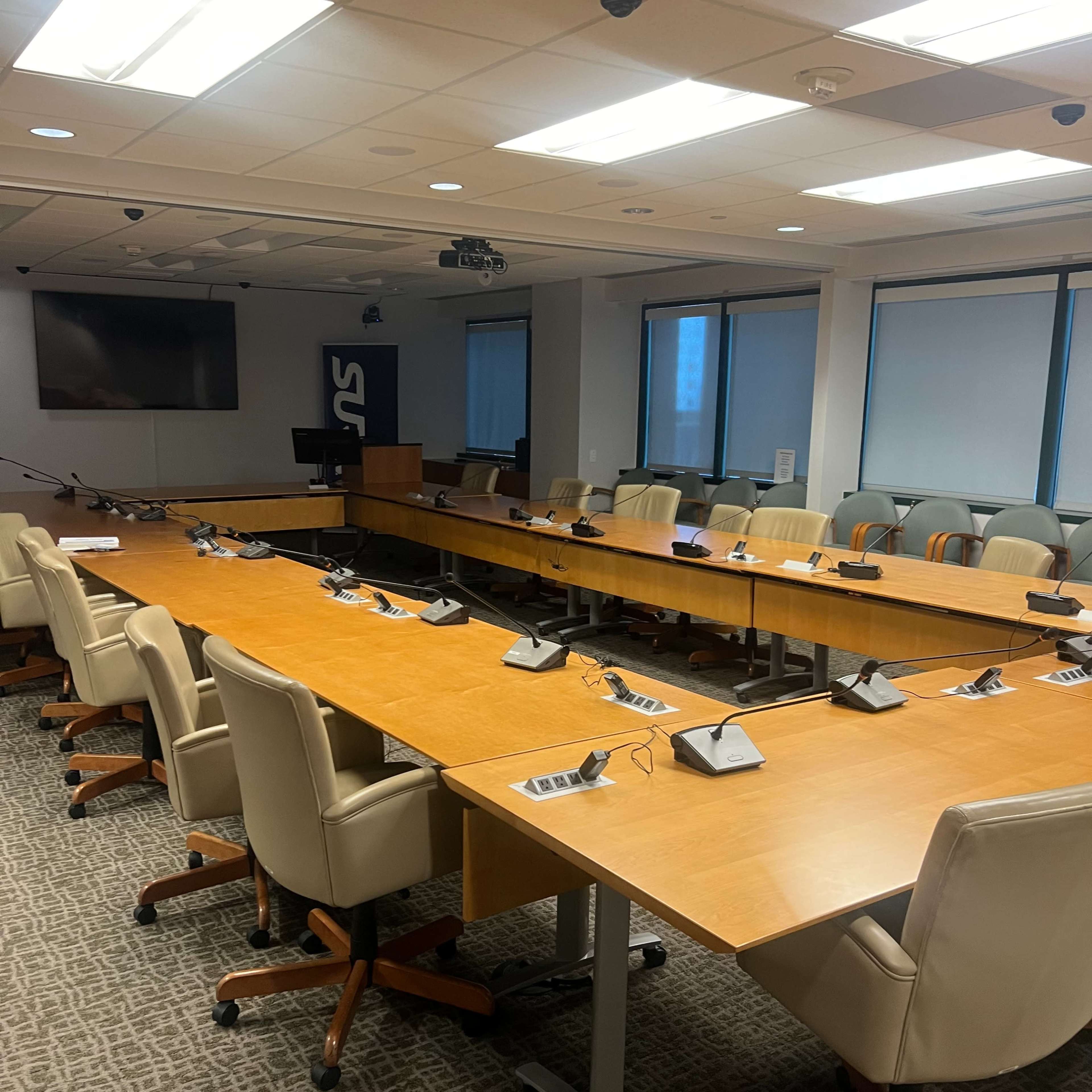 A large conference room features a long wooden table surrounded by office chairs and equipped with audio-visual technology.