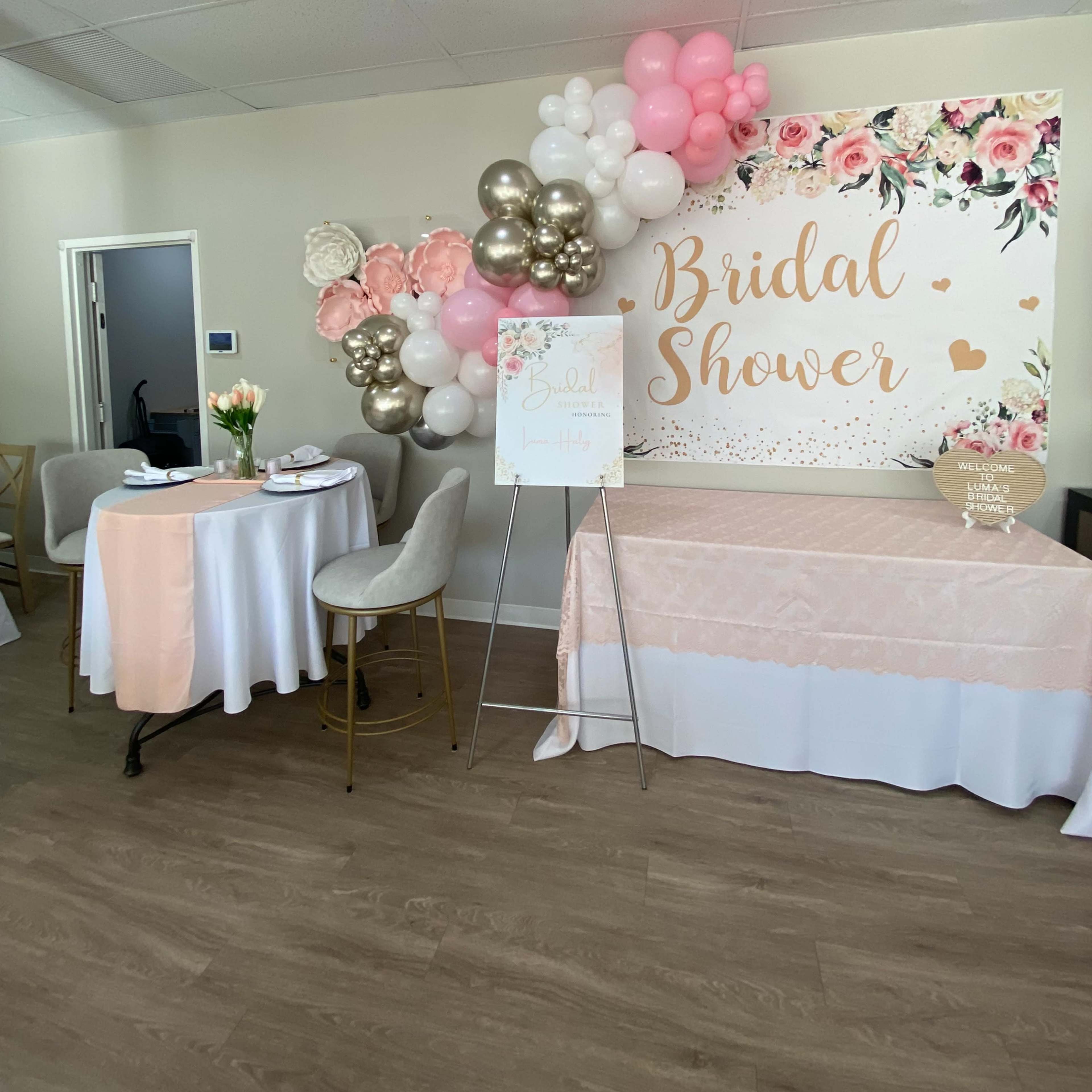 A decorated bridal shower setup featuring a table with a floral backdrop, a sign, balloons in pink and gold, and a small vase of flowers.