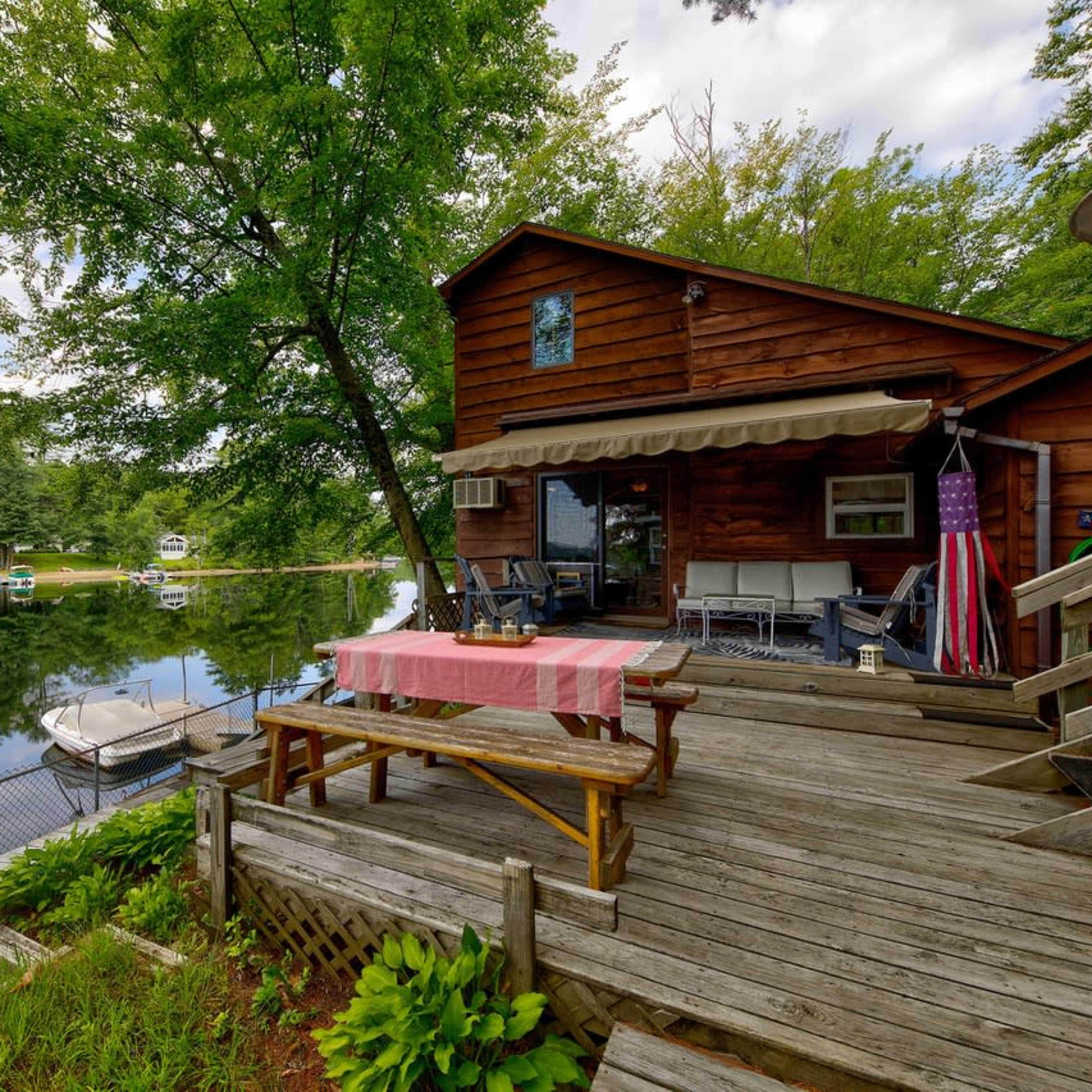 A wooden deck with a picnic table overlooks a calm lake surrounded by trees, with a house featuring a small awning in the background.