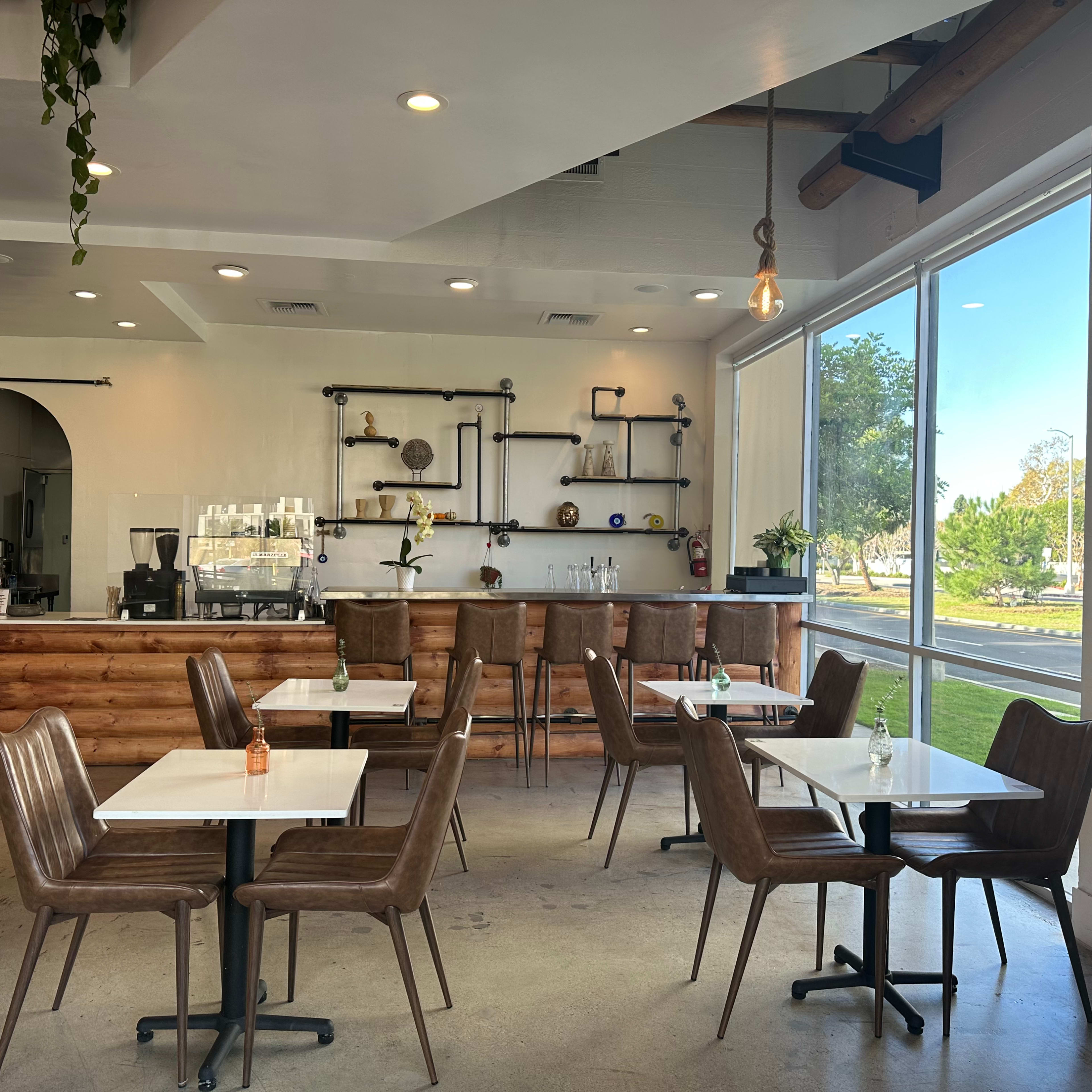 A modern café interior featuring wooden furniture, large windows, and a reception area with a counter and decorative items on the shelves.