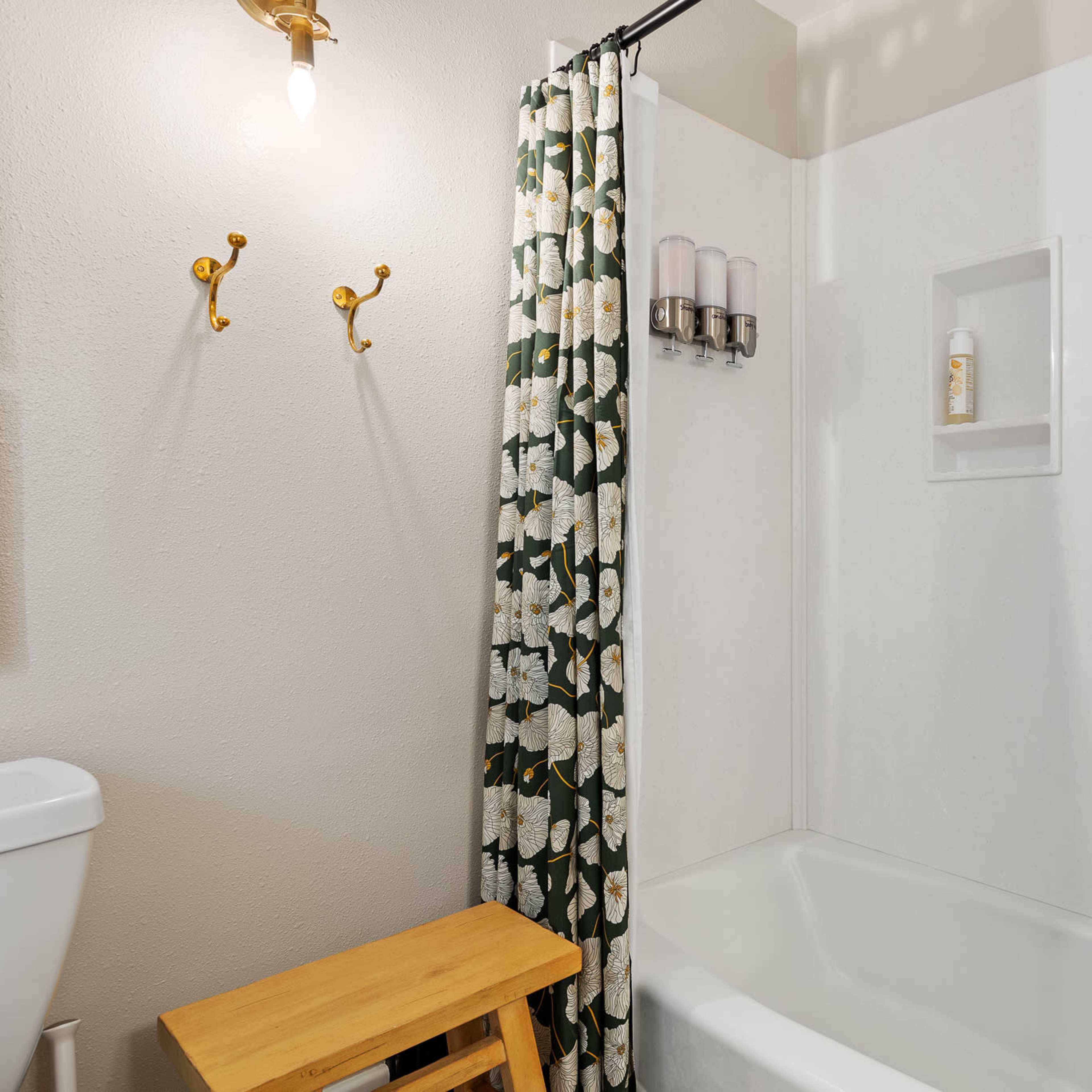 A bathroom with a shower-tub combination, a light fixture on the wall, a wooden stool, and a floral shower curtain.
