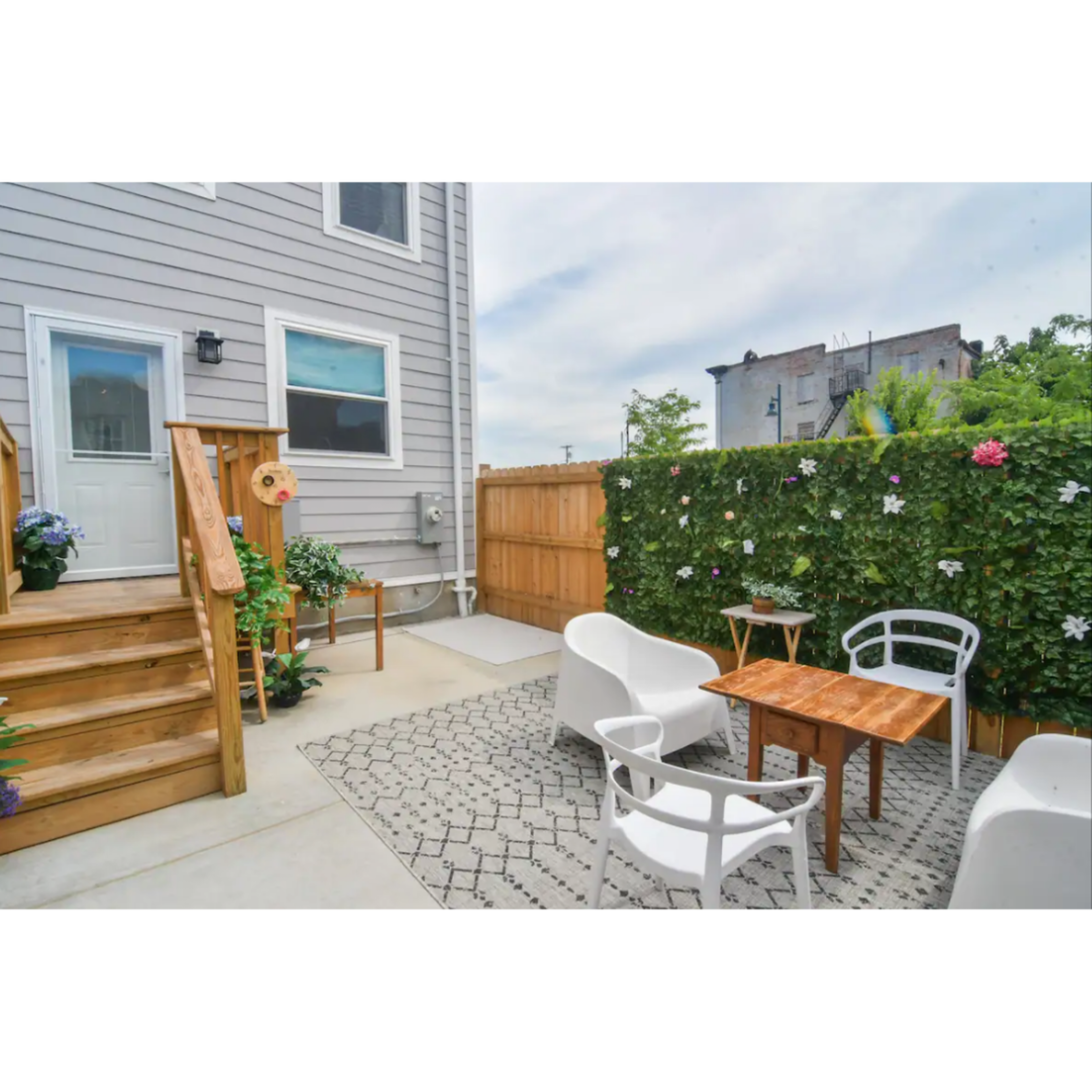 A small outdoor patio features a wooden table surrounded by white chairs, with steps leading up to a door and a wall covered in greenery.
