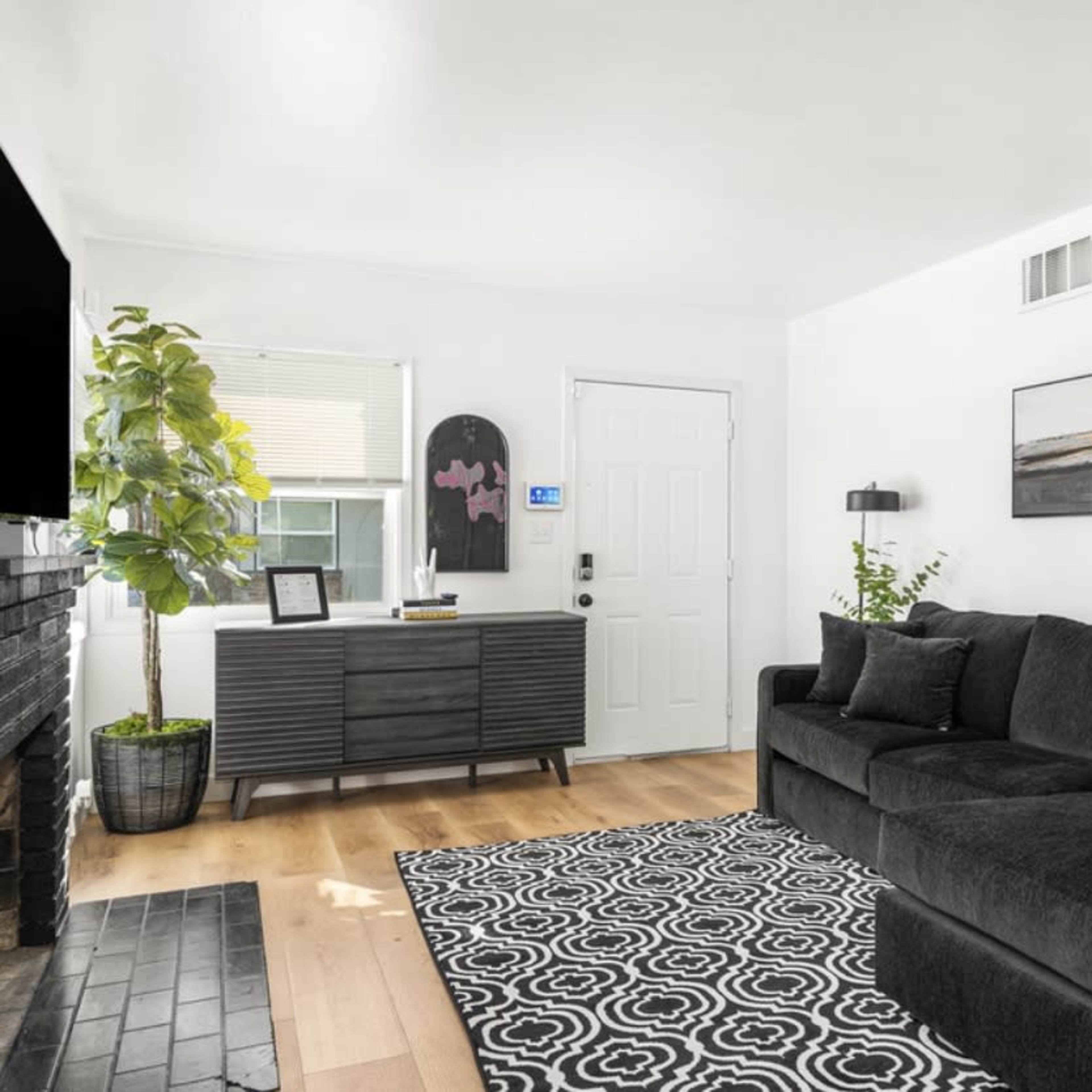 A modern living room features a dark sectional sofa, a black and white patterned rug, a console table, and a door leading outside.