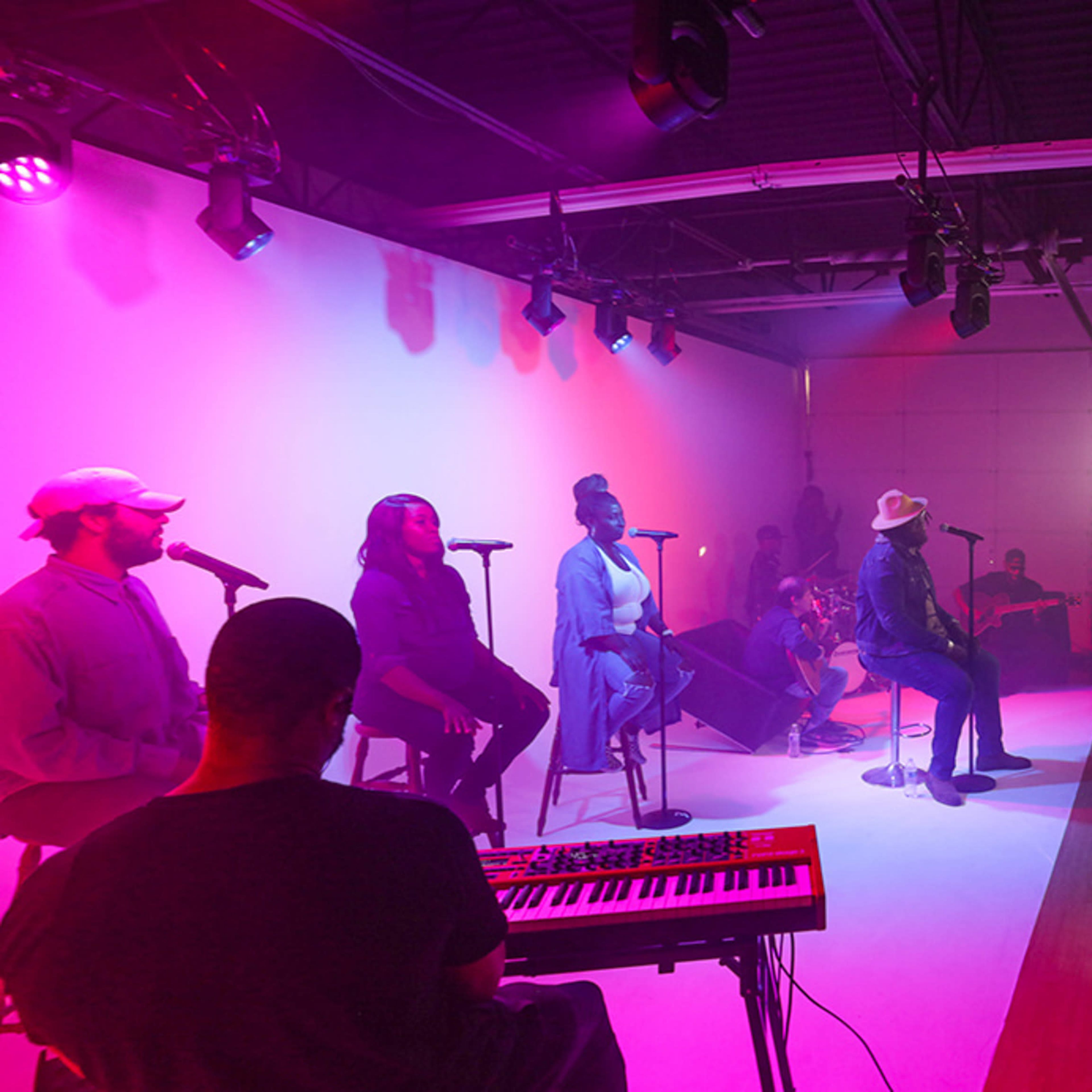 A group of musicians performs on stage under colorful lighting while a technician operates equipment nearby.
