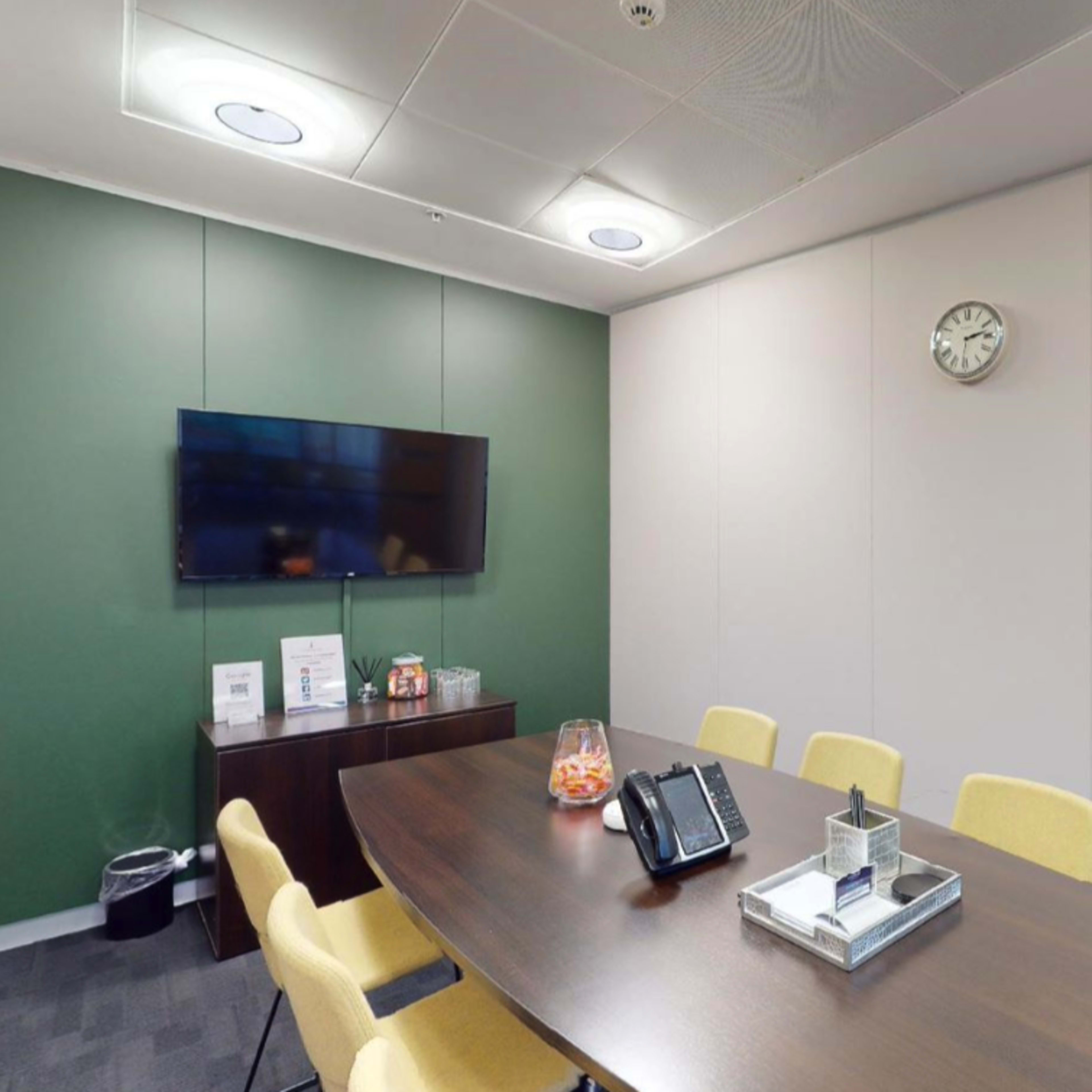 A conference room features a table with yellow chairs, a television mounted on the wall, and a clock above on a plain backdrop.