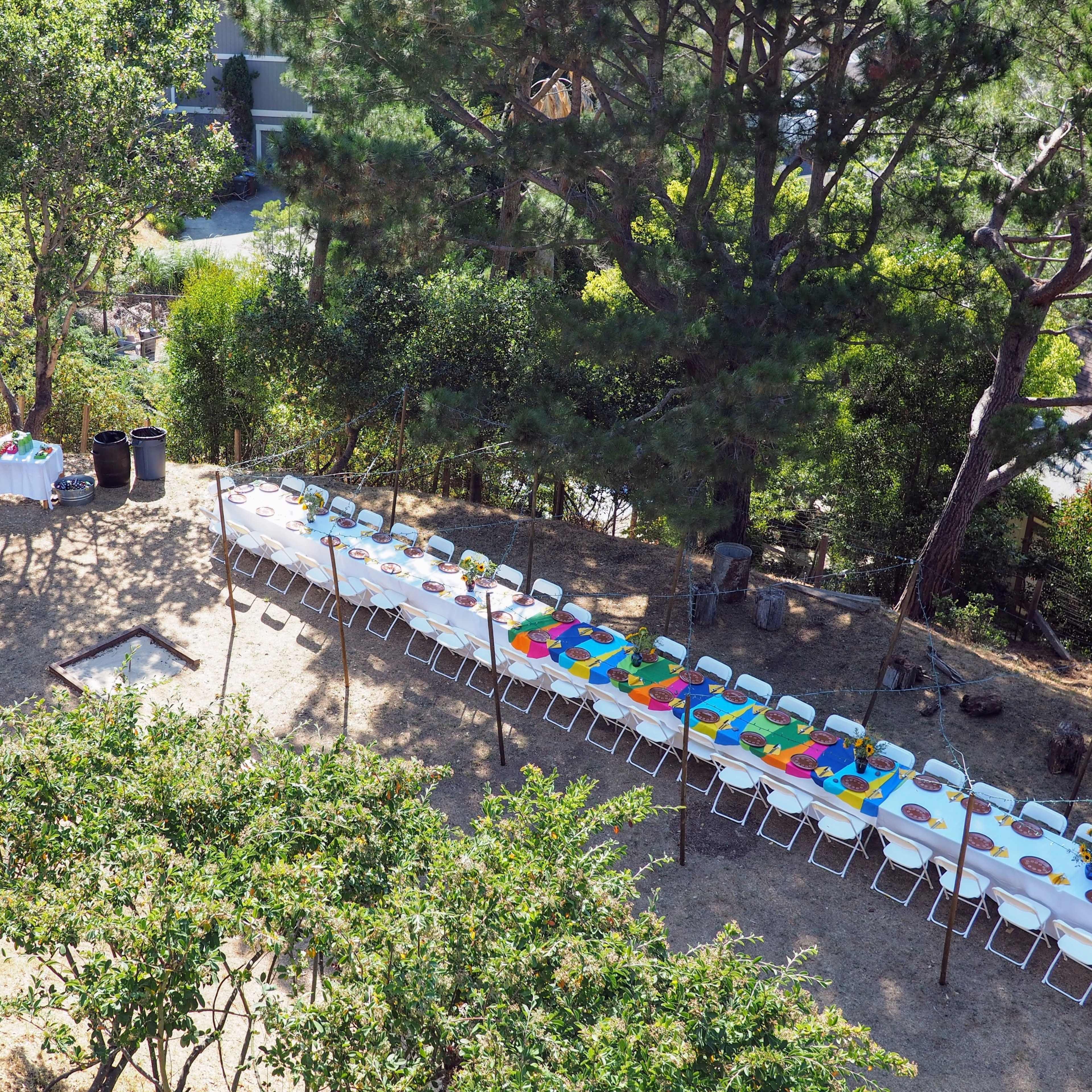 A long table is set under trees for an outdoor gathering, with two individuals arranging items nearby.