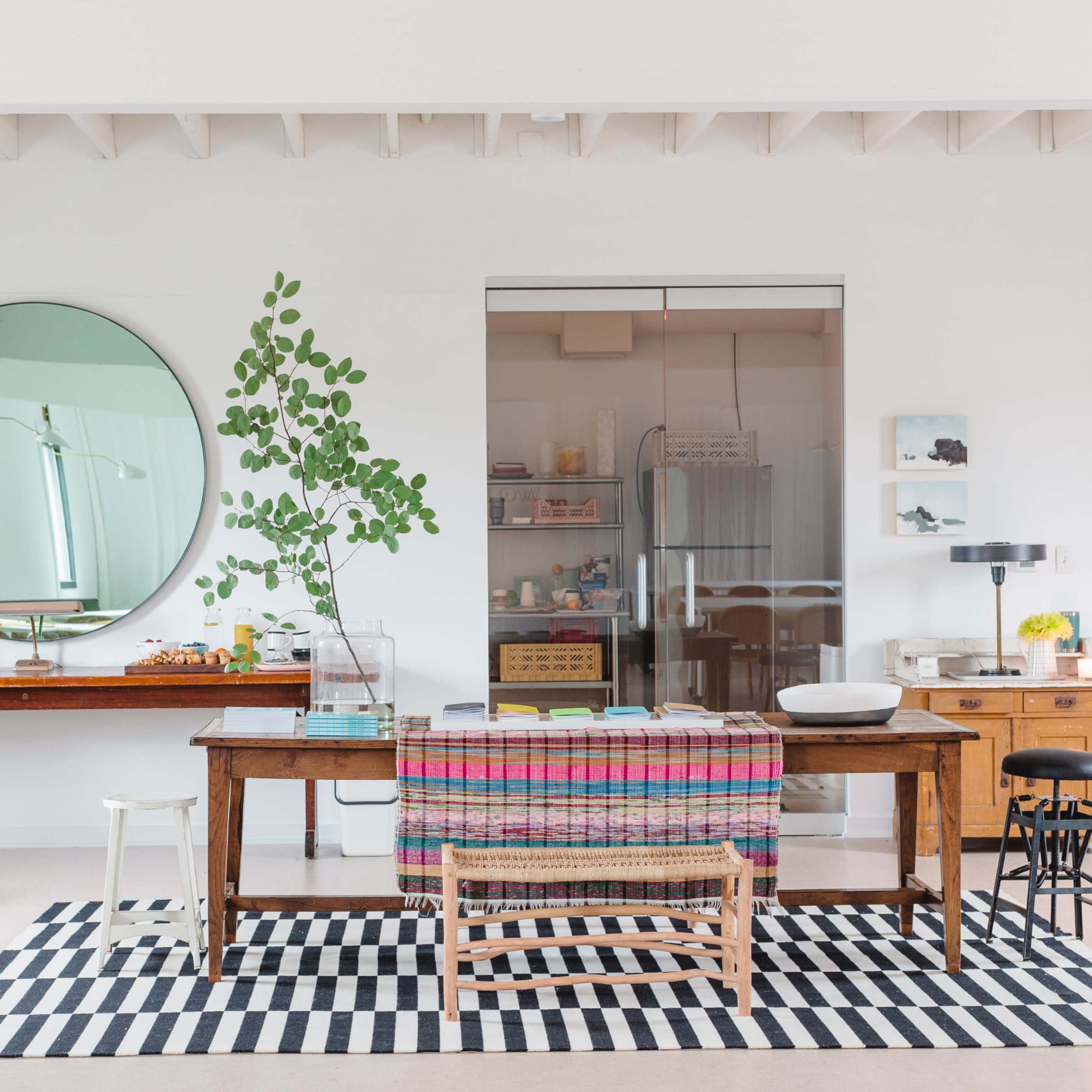 A modern workspace featuring a wooden table draped with a colorful throw, surrounded by various plants, a round mirror, and a glass partition separating a kitchen area.