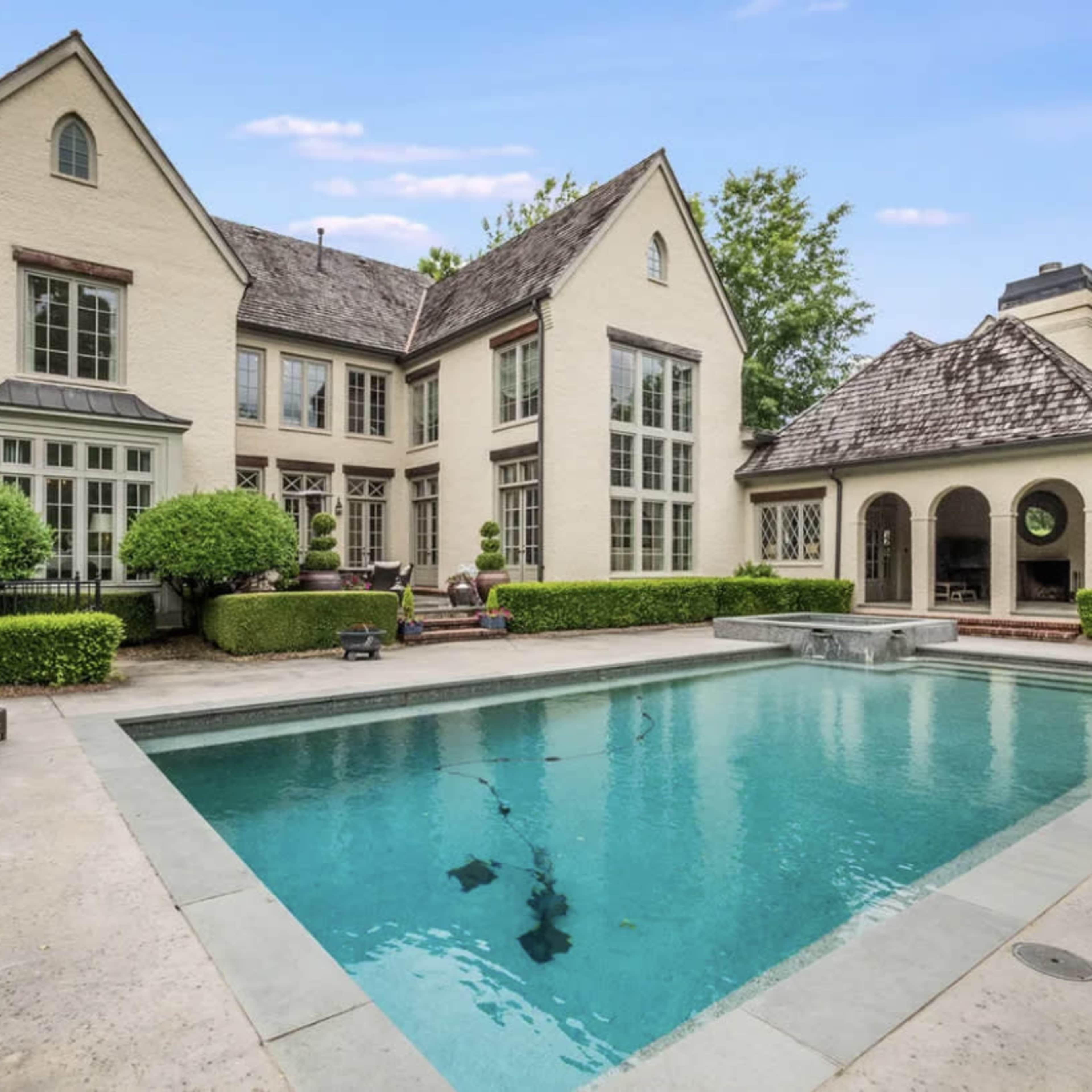 A large house with a pool in the backyard, surrounded by neatly trimmed hedges and a separate structure that appears to be a patio or gazebo.