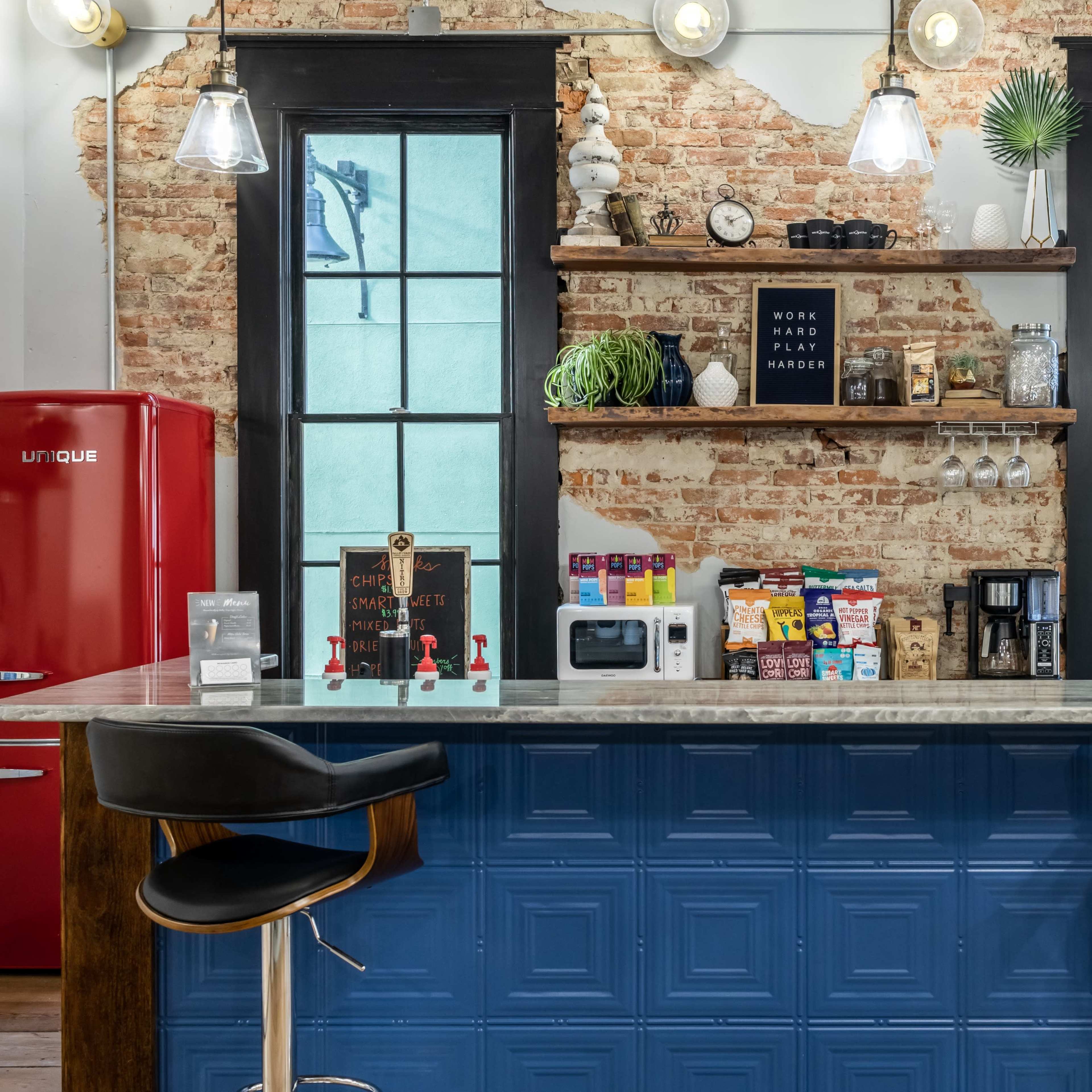 A modern kitchen features a red refrigerator, a granite countertop with bar stools, and shelves displaying various kitchen items against a brick wall.