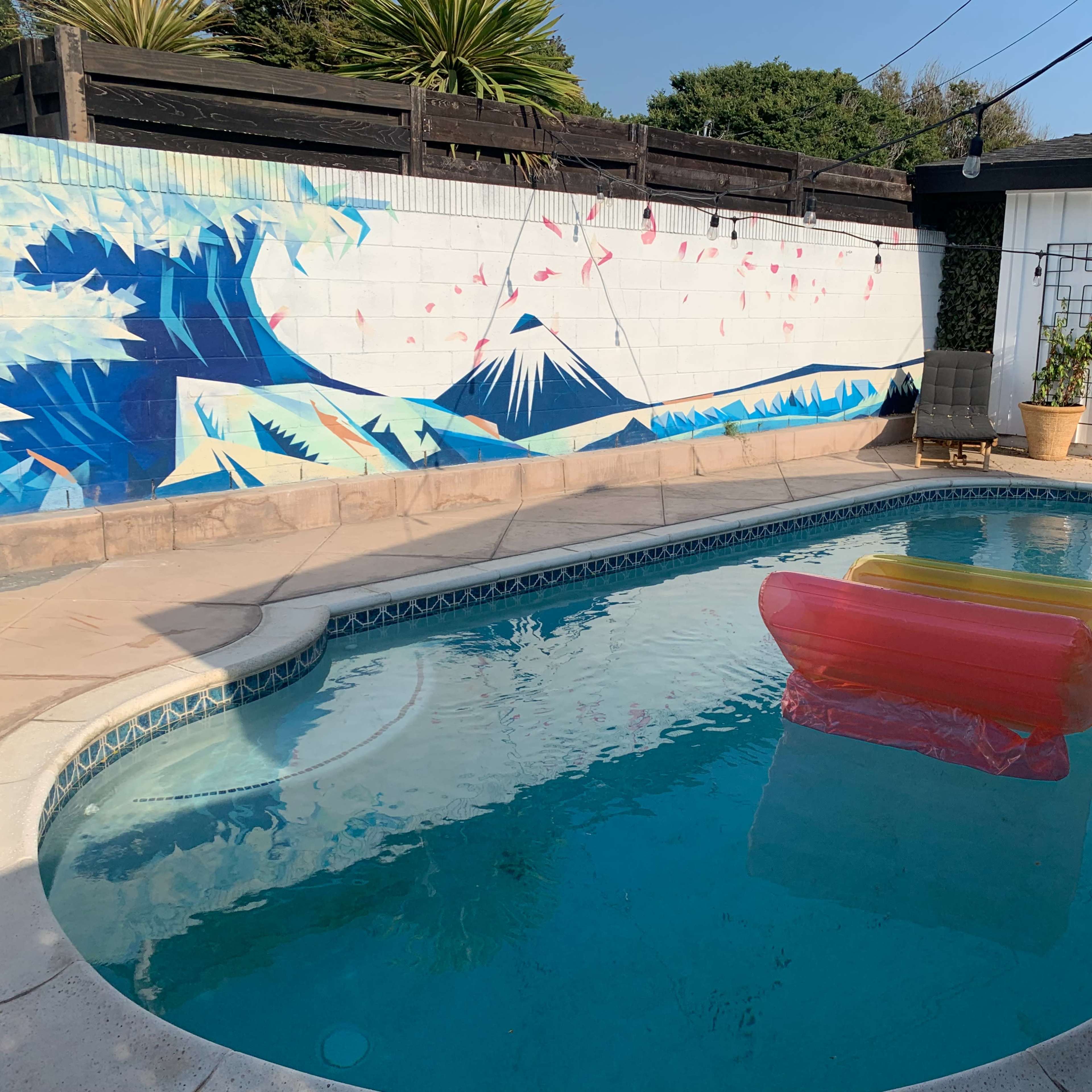 The scene features a swimming pool with a pink and yellow inflatable float, surrounded by a mural of waves and a mountain on the wall.