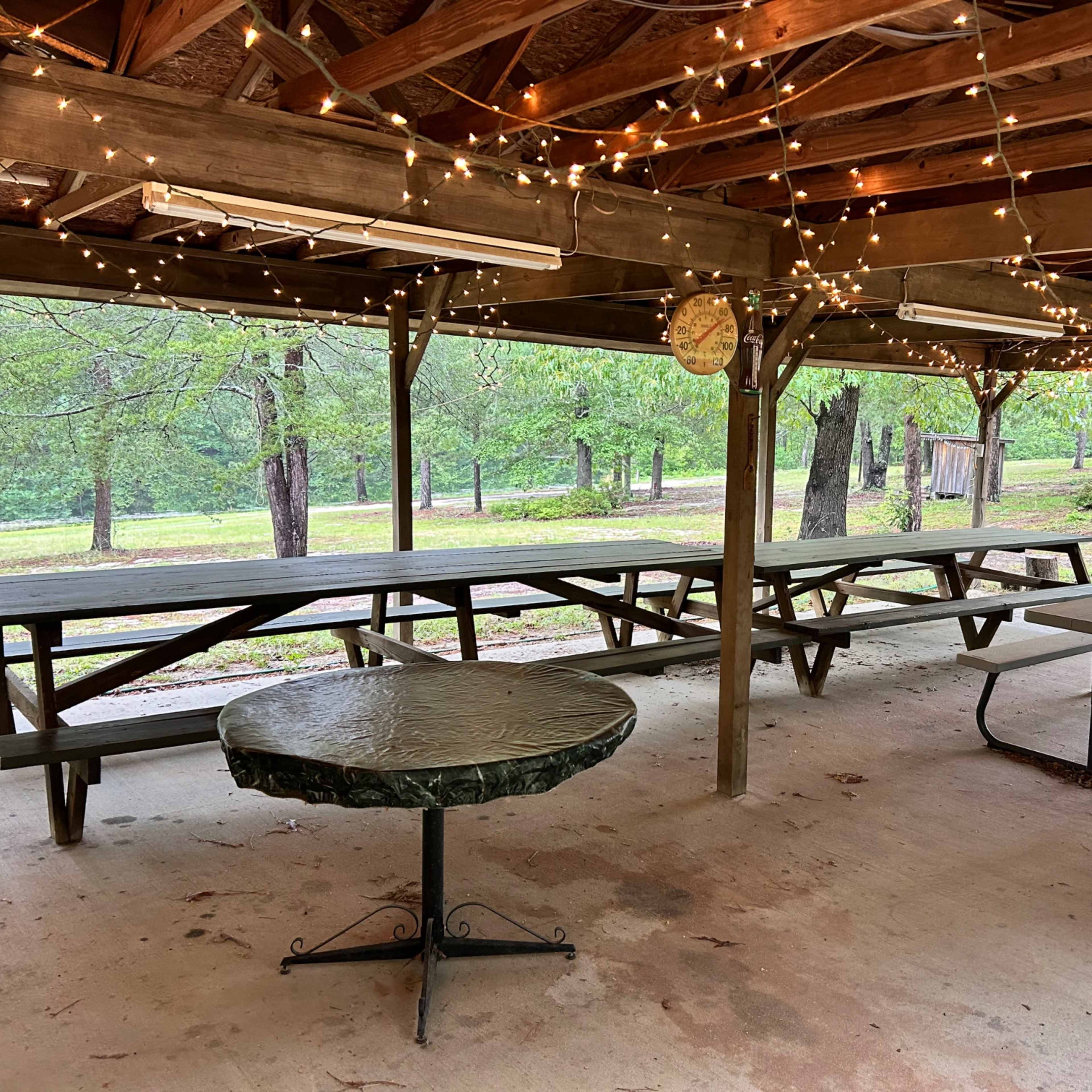 An open pavilion with wooden beams and string lights, featuring several picnic tables and a round table underneath.