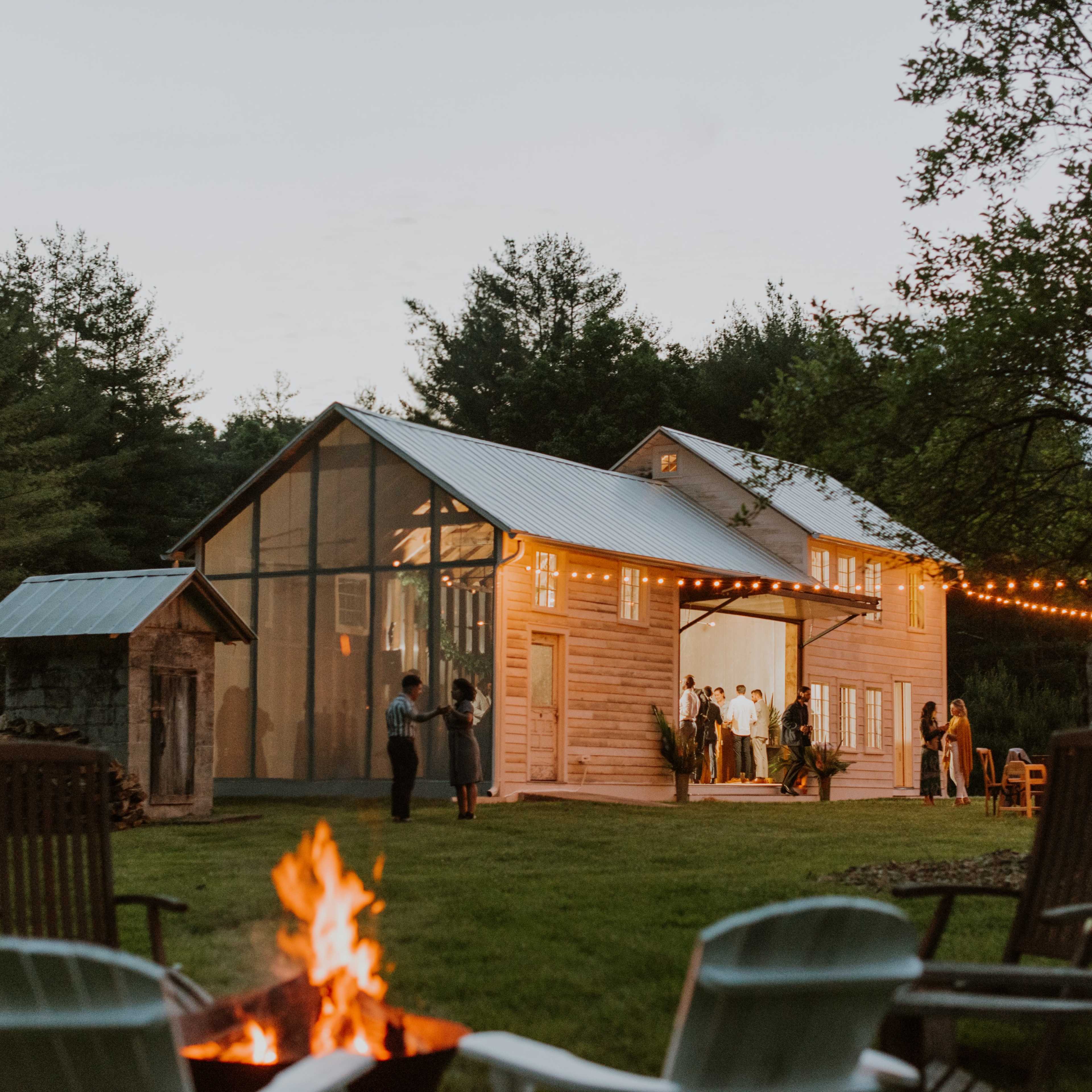 A wooden barn is illuminated in the evening with guests gathered outside around a fire pit and string lights overhead.
