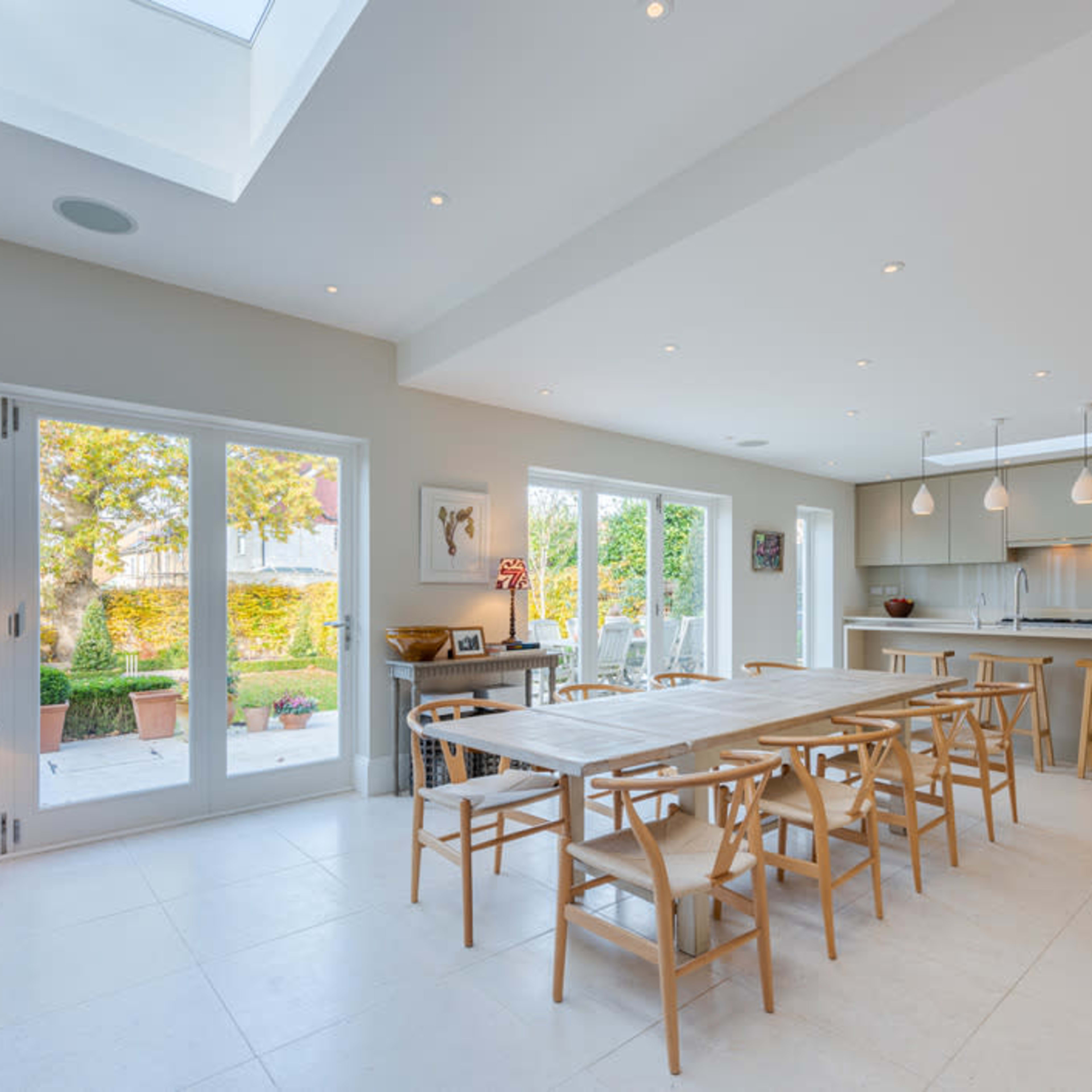 A modern kitchen and dining area with a large table, wooden stools, and glass doors opening to a garden.