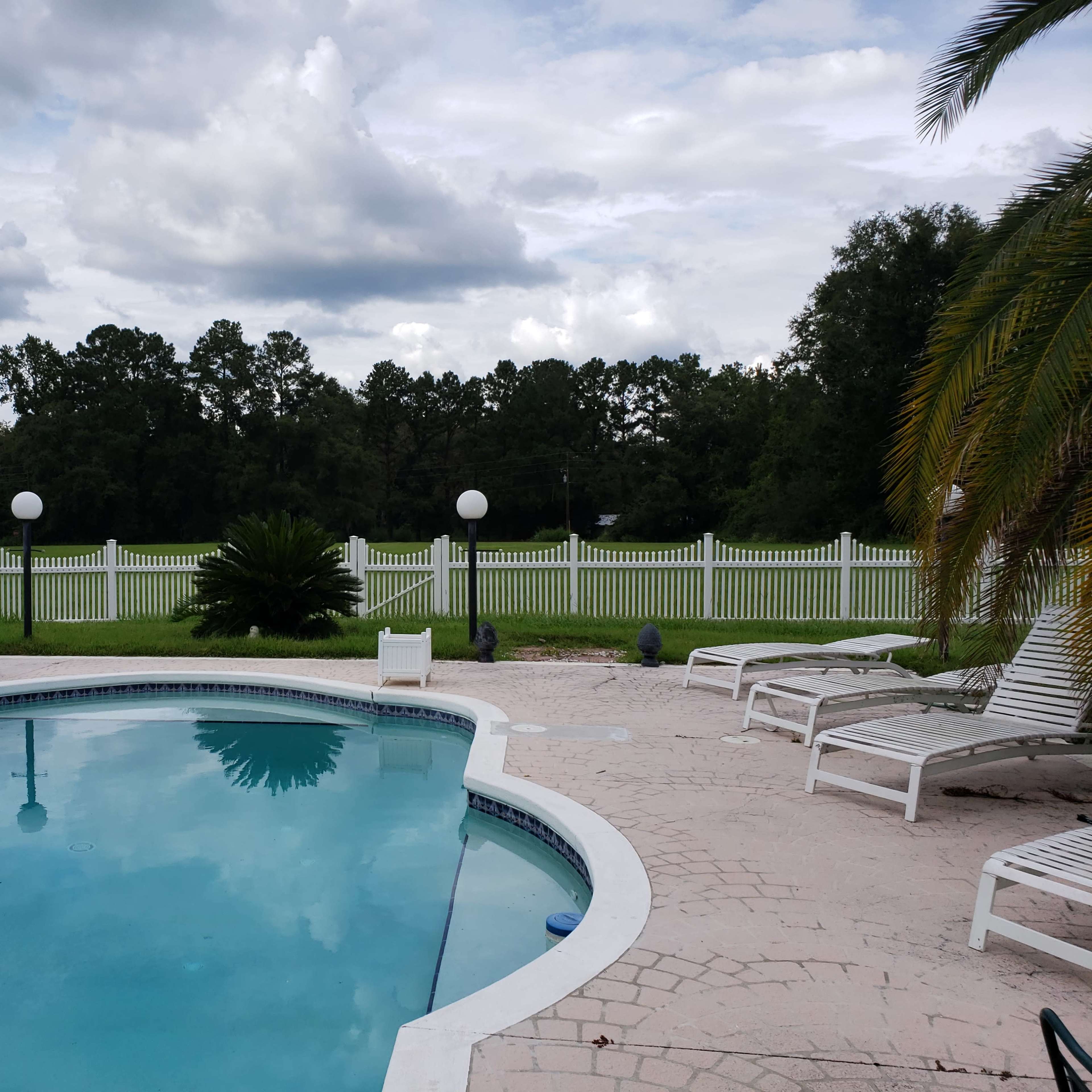 A clear swimming pool is surrounded by lounge chairs and a white fence, with trees and a cloudy sky in the background.