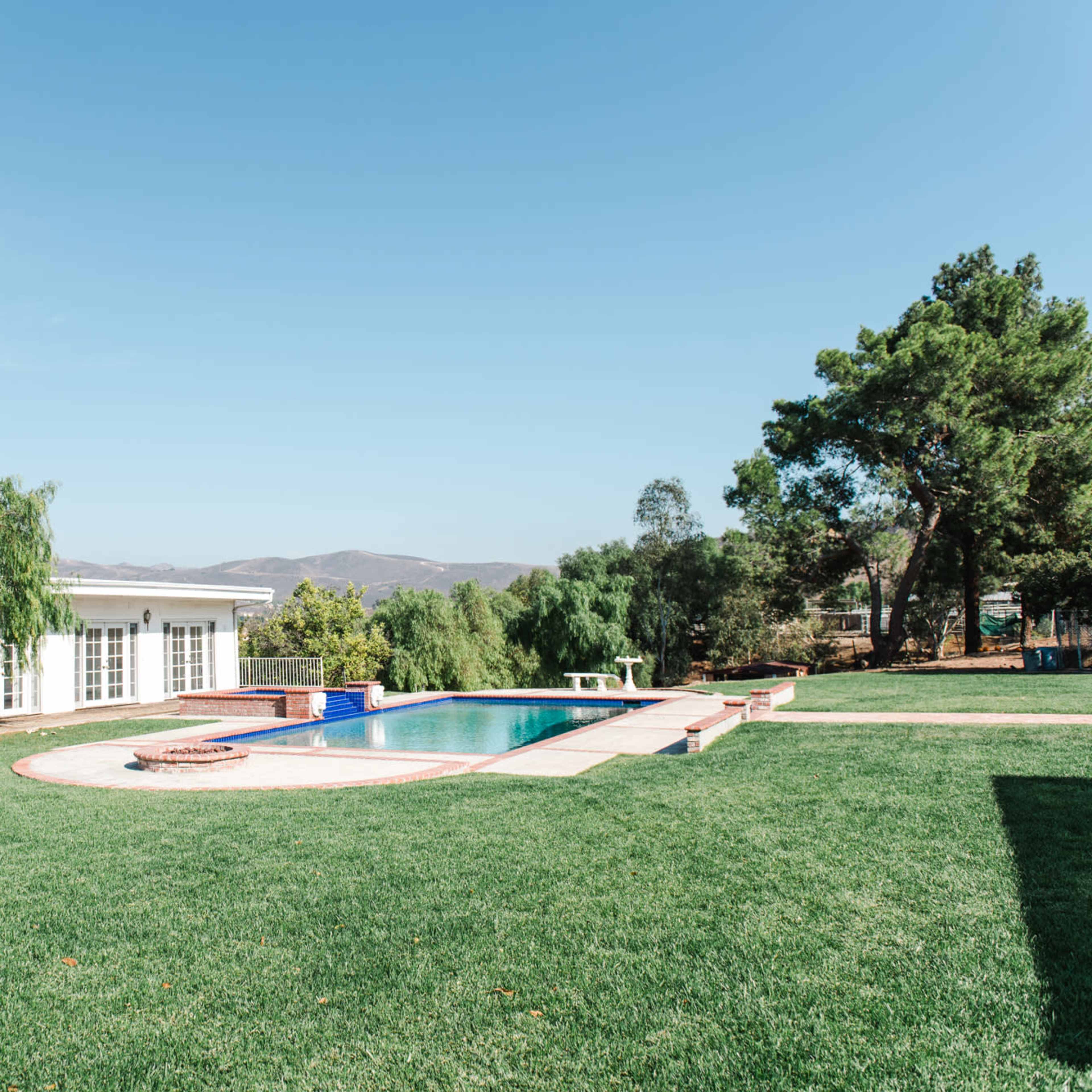 A modern home with a swimming pool is surrounded by landscaped grass and trees under a clear blue sky.
