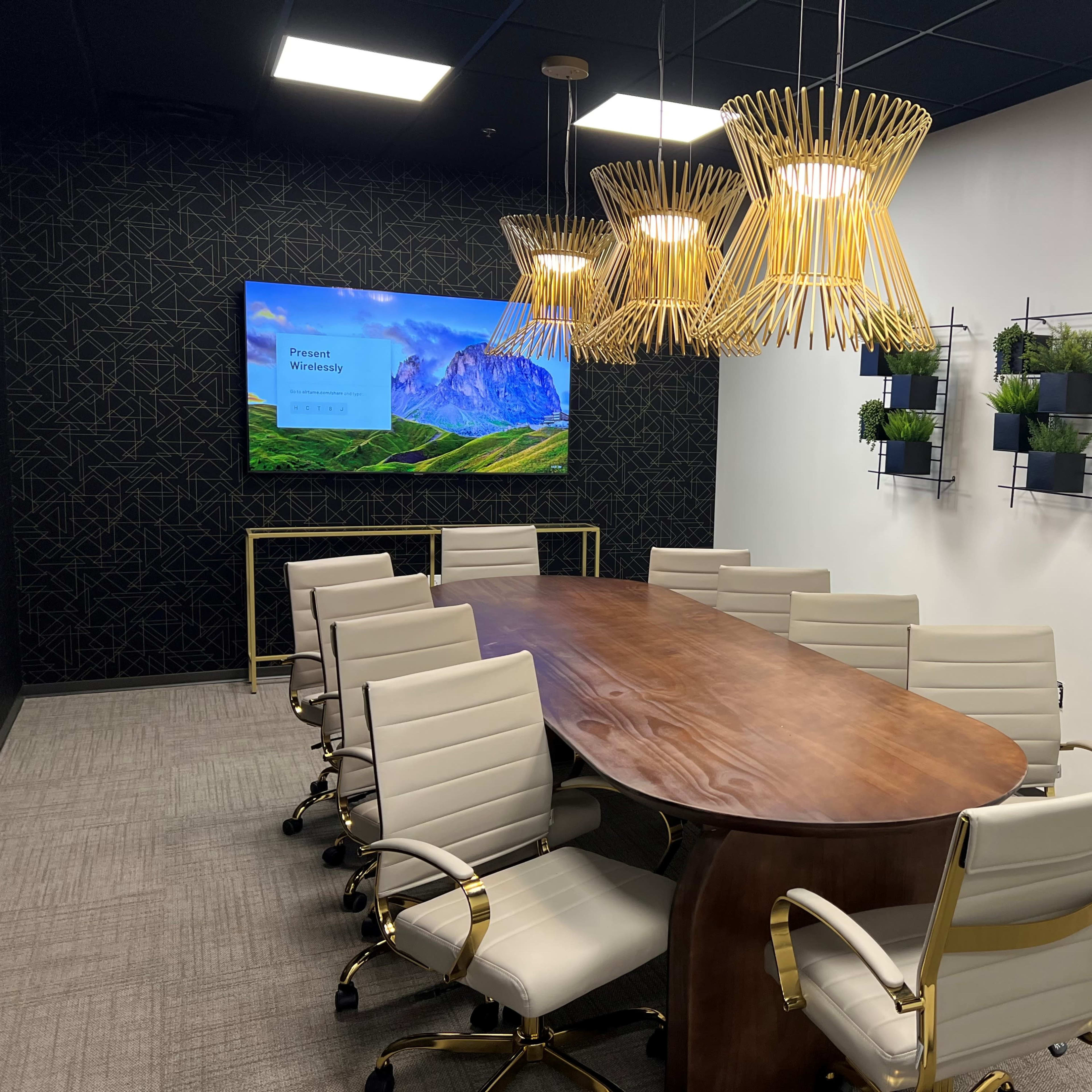 A modern conference room featuring a long wooden table, white chairs, a wall-mounted screen displaying a scenic landscape, and green plants arranged on wall shelves.