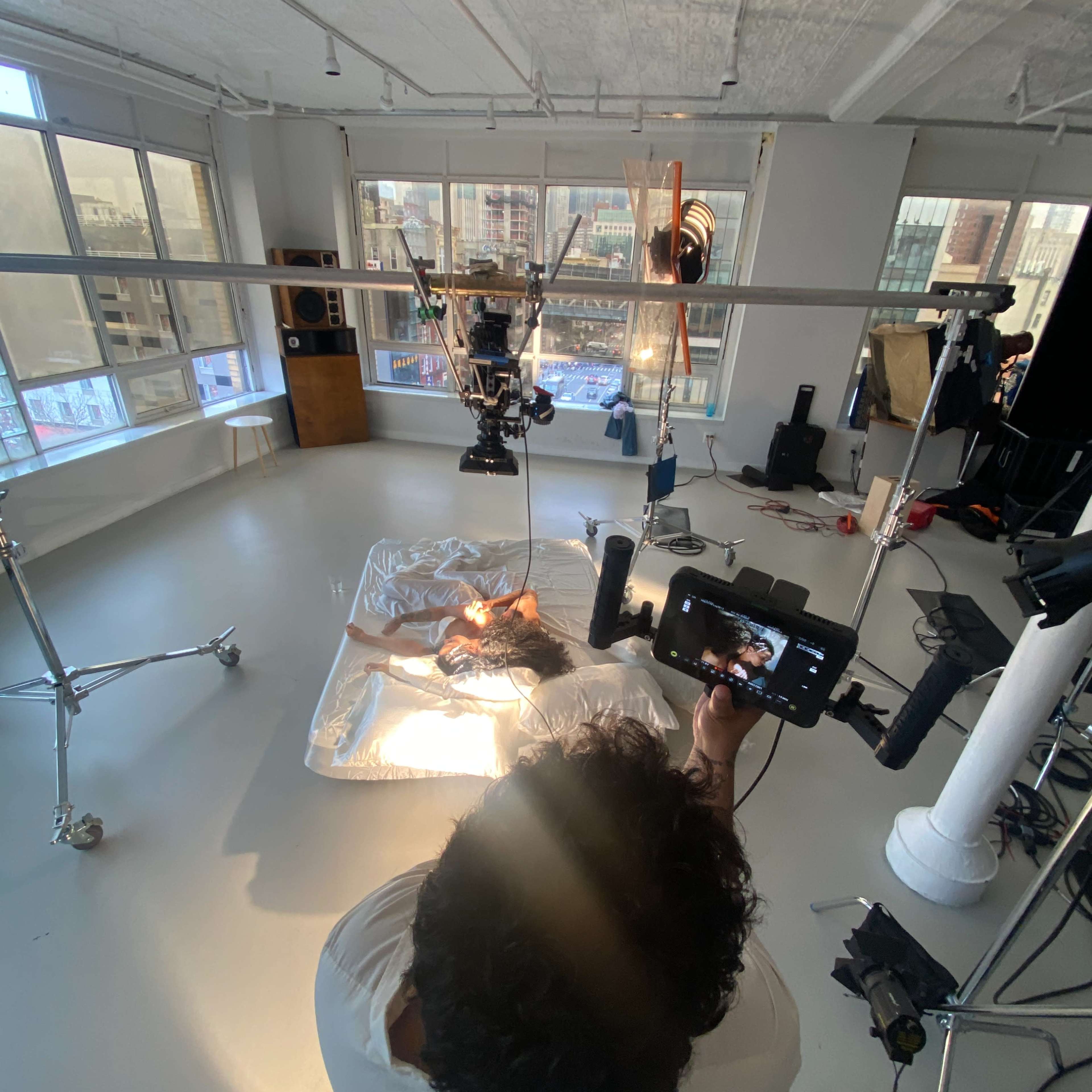 A person operates a camera from above a setup with a reflective surface and a subject in a studio filled with various equipment and large windows.