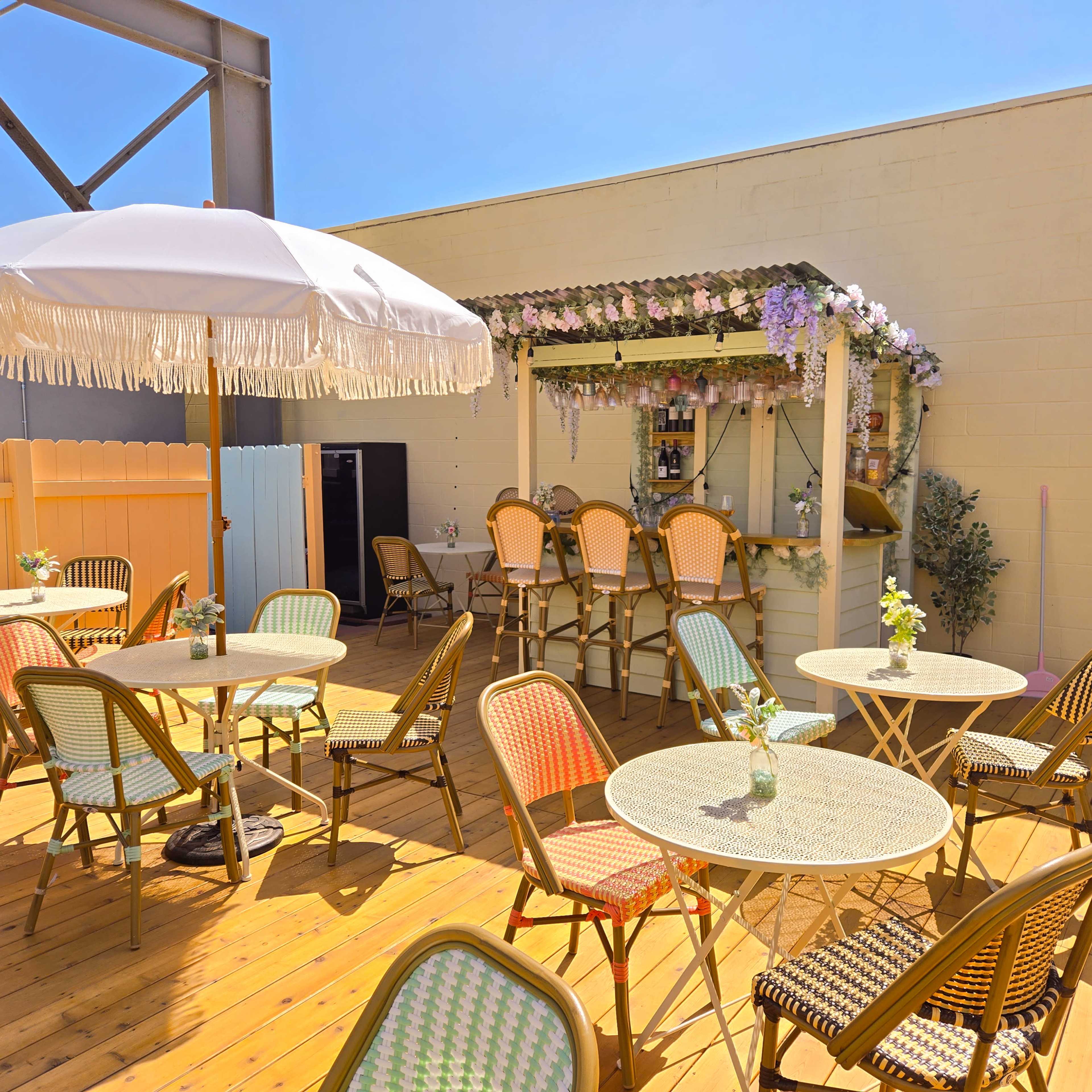 A sunny outdoor patio features colorful bistro tables and chairs arranged around a small bar decorated with flowers.