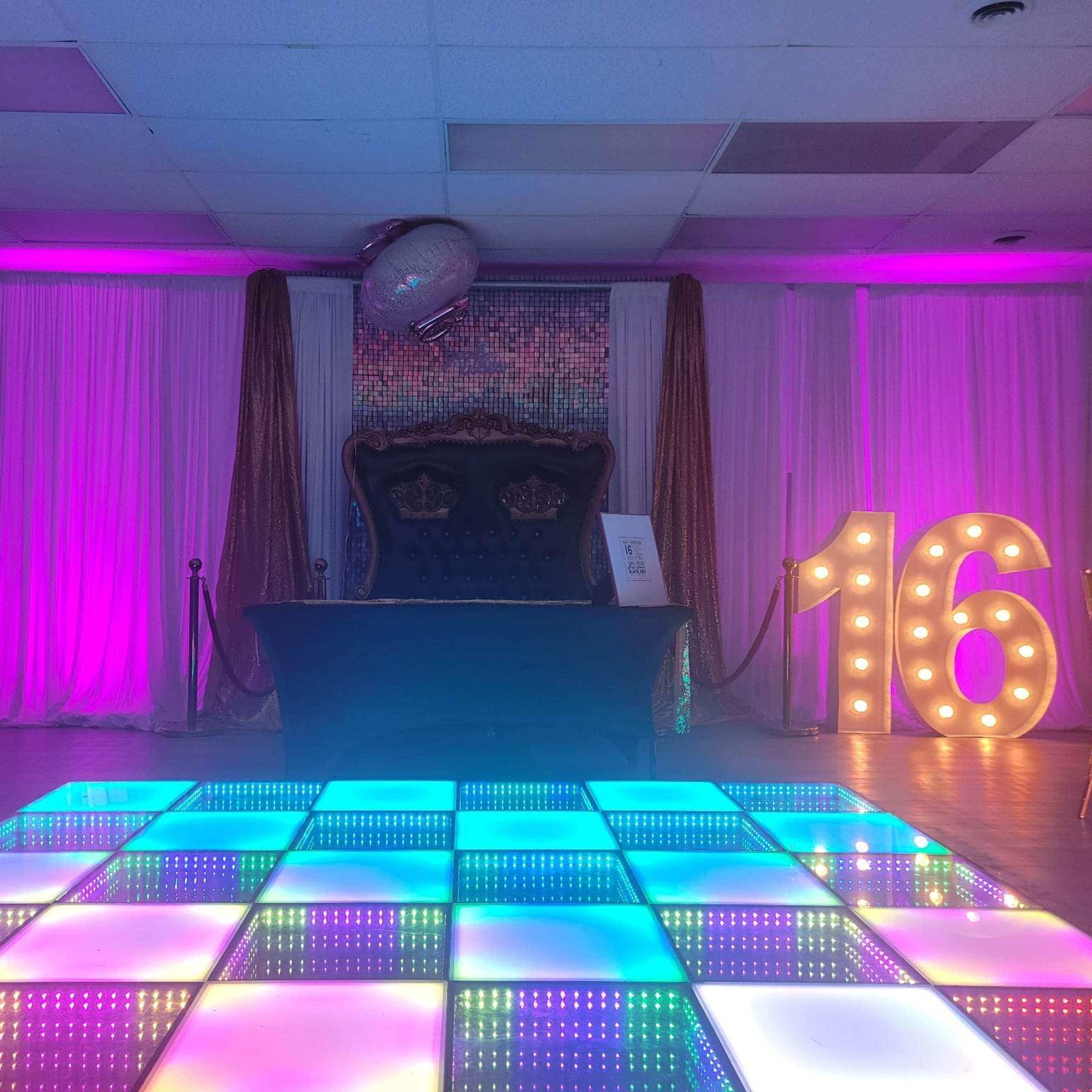 A decorated event space featuring a light-up dance floor, a large illuminated "16" sign, and a central table with a backdrop of draped fabric and colored lights.