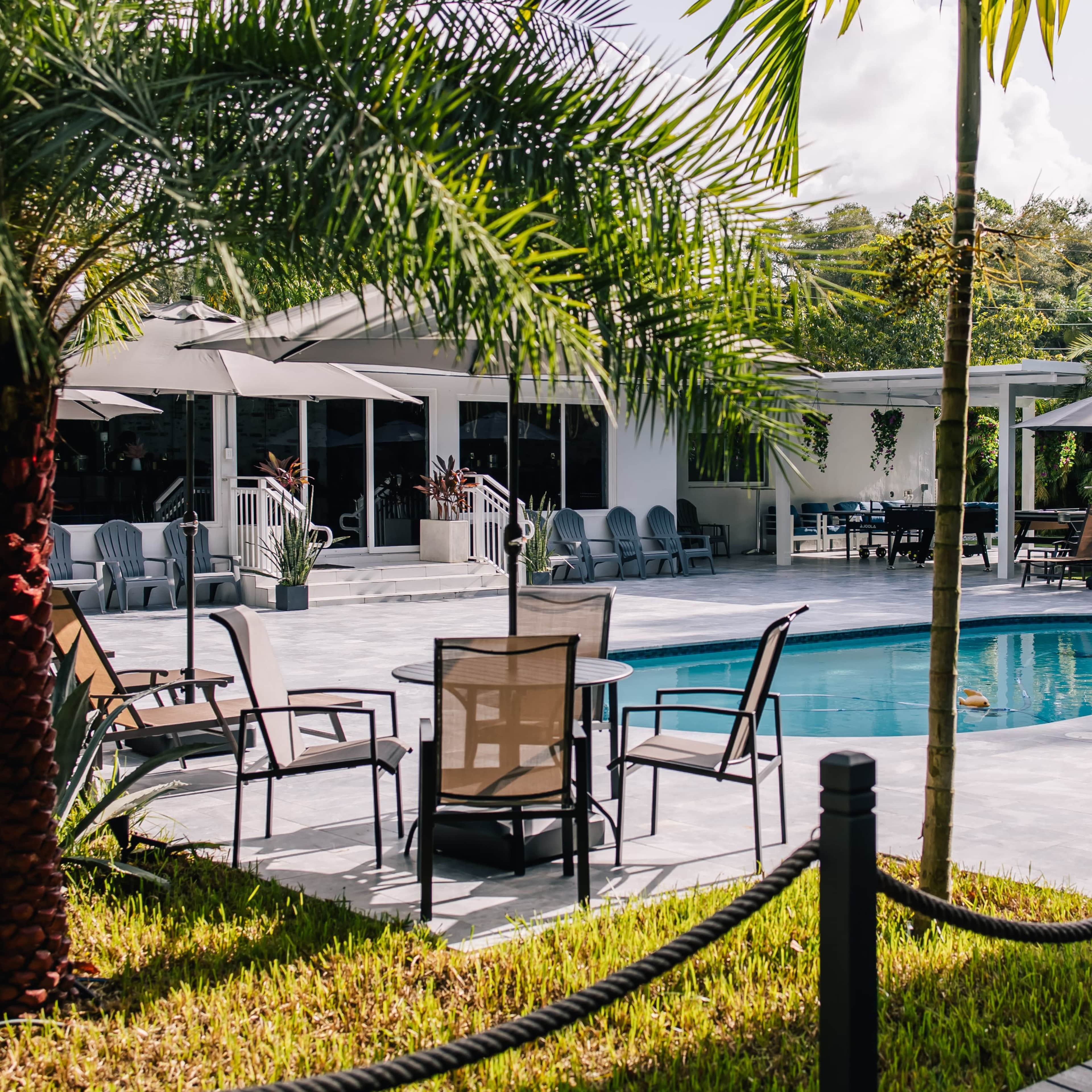 A swimming pool is surrounded by chairs and tables, with palm trees and umbrellas providing shade in a landscaped outdoor area.