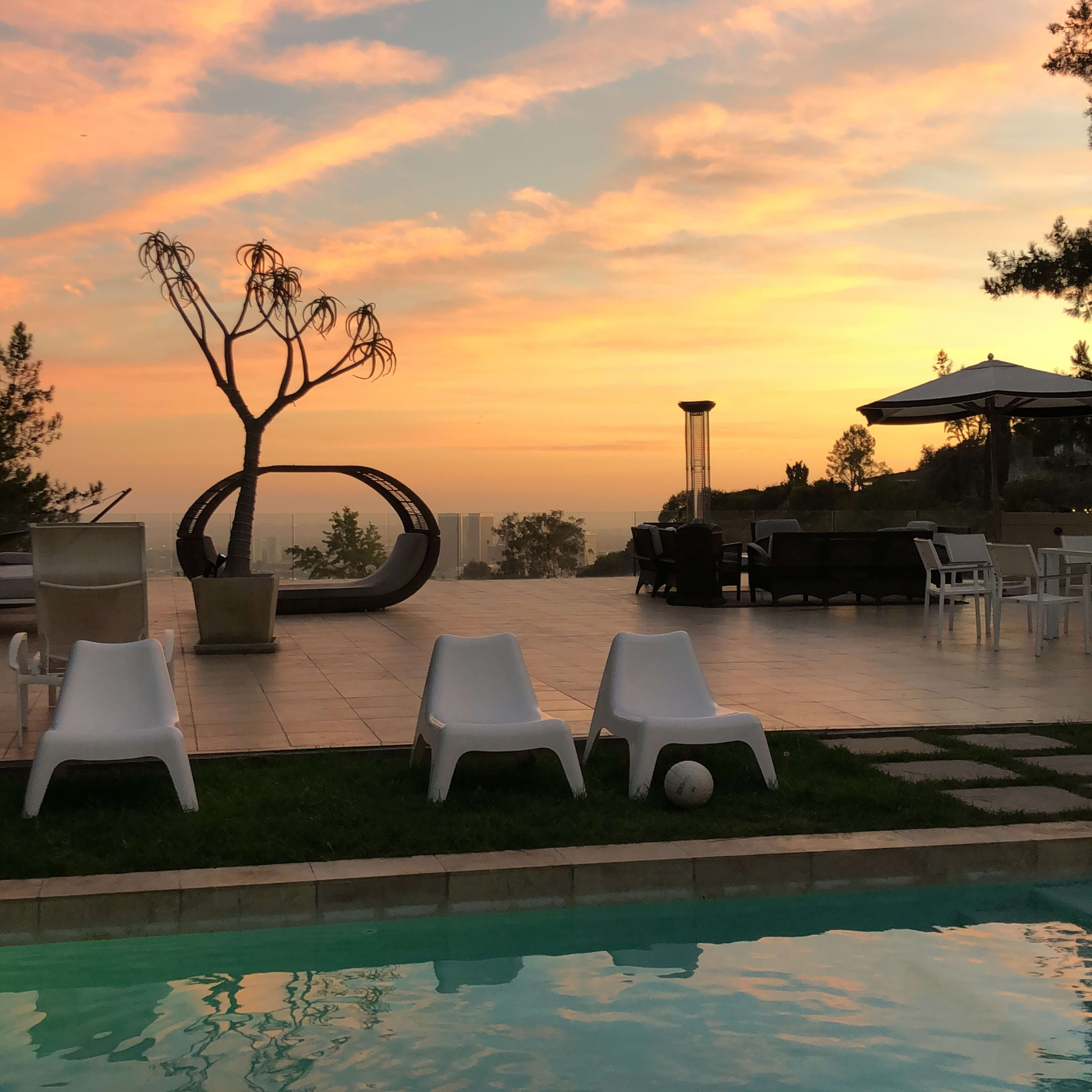 A poolside area with white chairs and a tree silhouetted against a sunset sky filled with clouds.